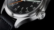 Close-up of a stainless steel watch case with a black dial, orange second hand, and black leather strap.