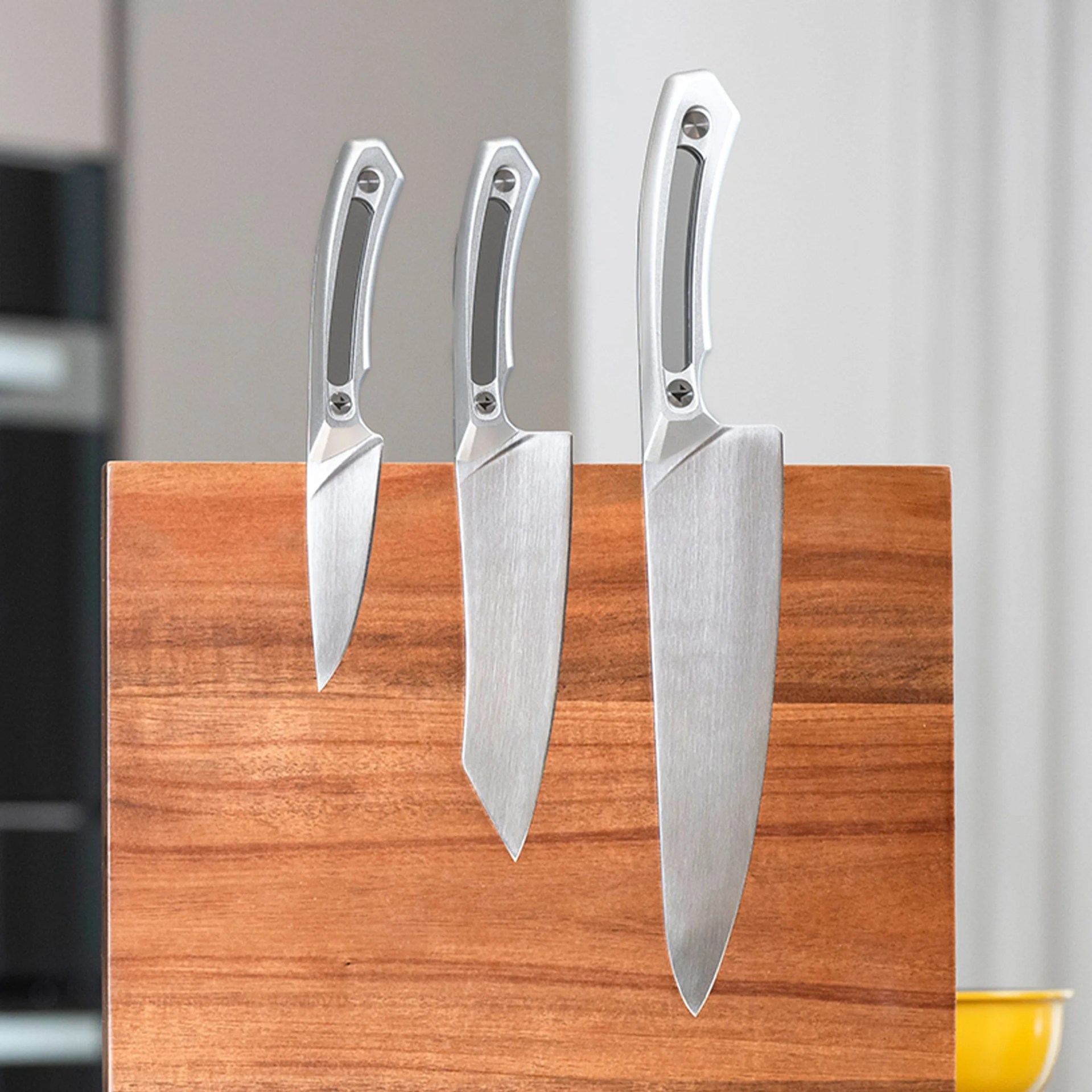 Three stainless steel kitchen knives with silver handles attached to a wooden magnetic knife block.