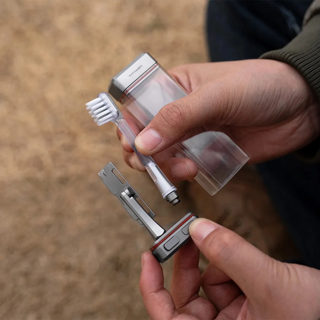 Hands holding a compact titanium travel toothbrush with a transparent case and metal base.
