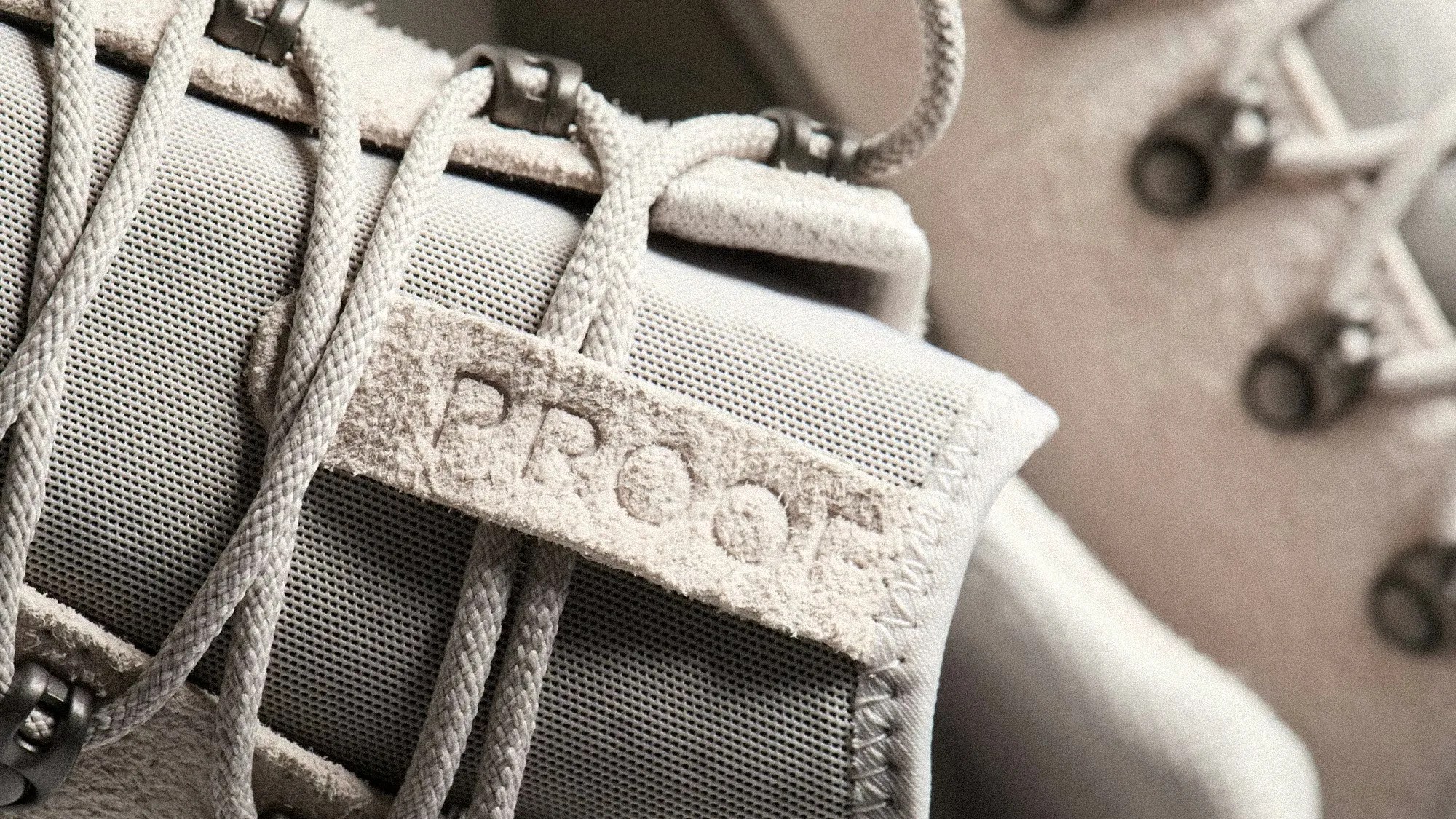 Close-up of beige suede tag with "PROOF" embossed on a light gray mesh fabric with beige laces threaded through metal eyelets.
