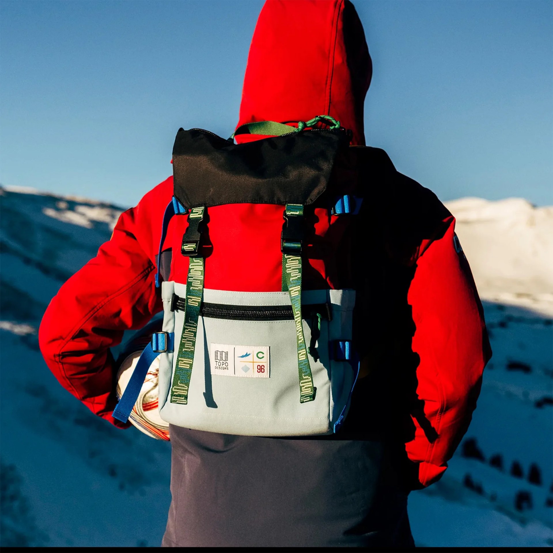 Red, black, and gray Topo Designs backpack with green straps worn by a person in a red jacket in a snowy mountain setting.