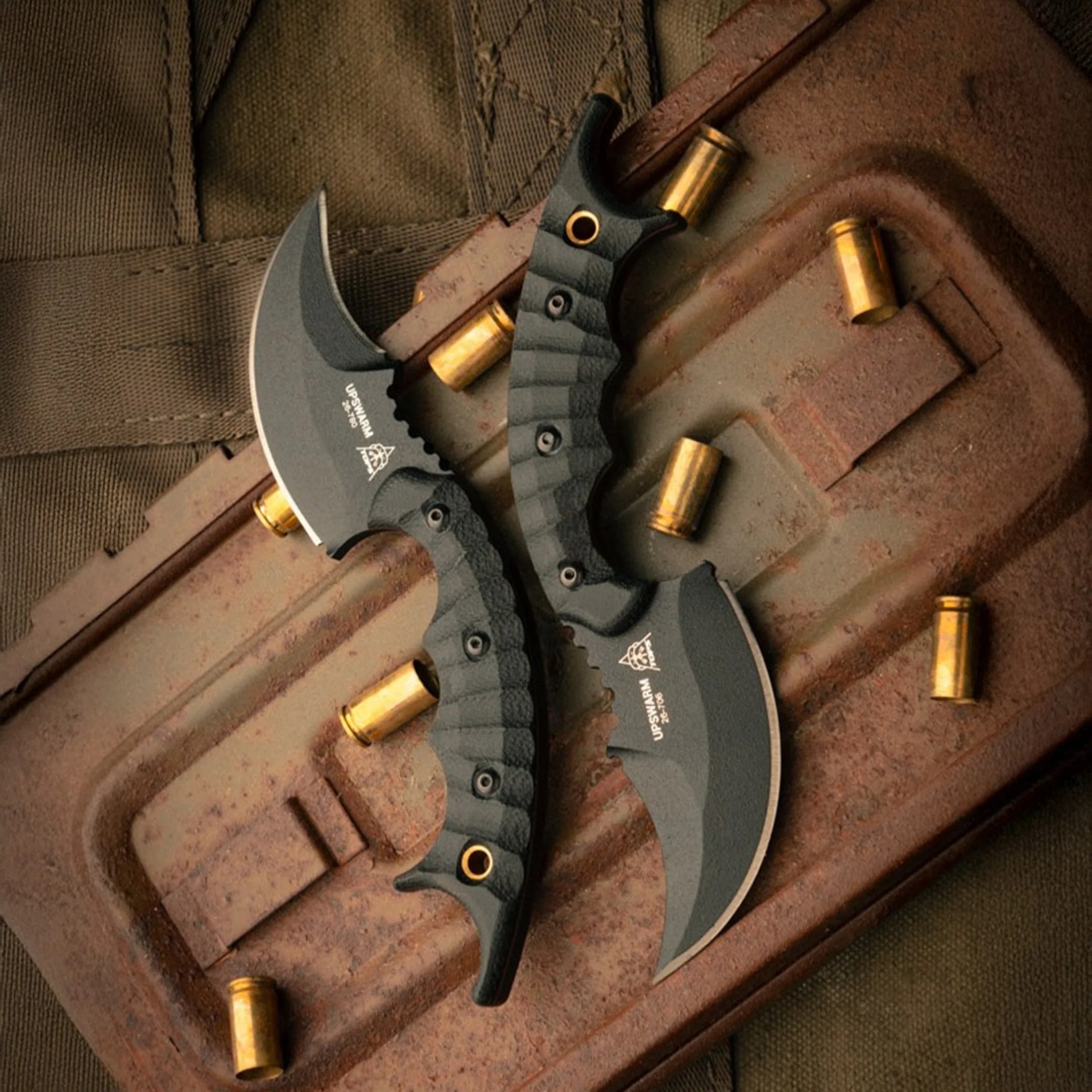 Two black curved karambit knives with textured handles on a rusty metal surface surrounded by brass bullet casings.