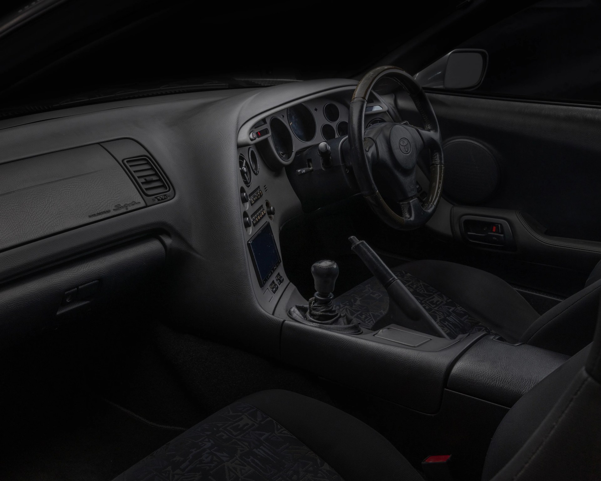 Black interior of a Toyota Supra with a manual gear shift and patterned fabric seats.