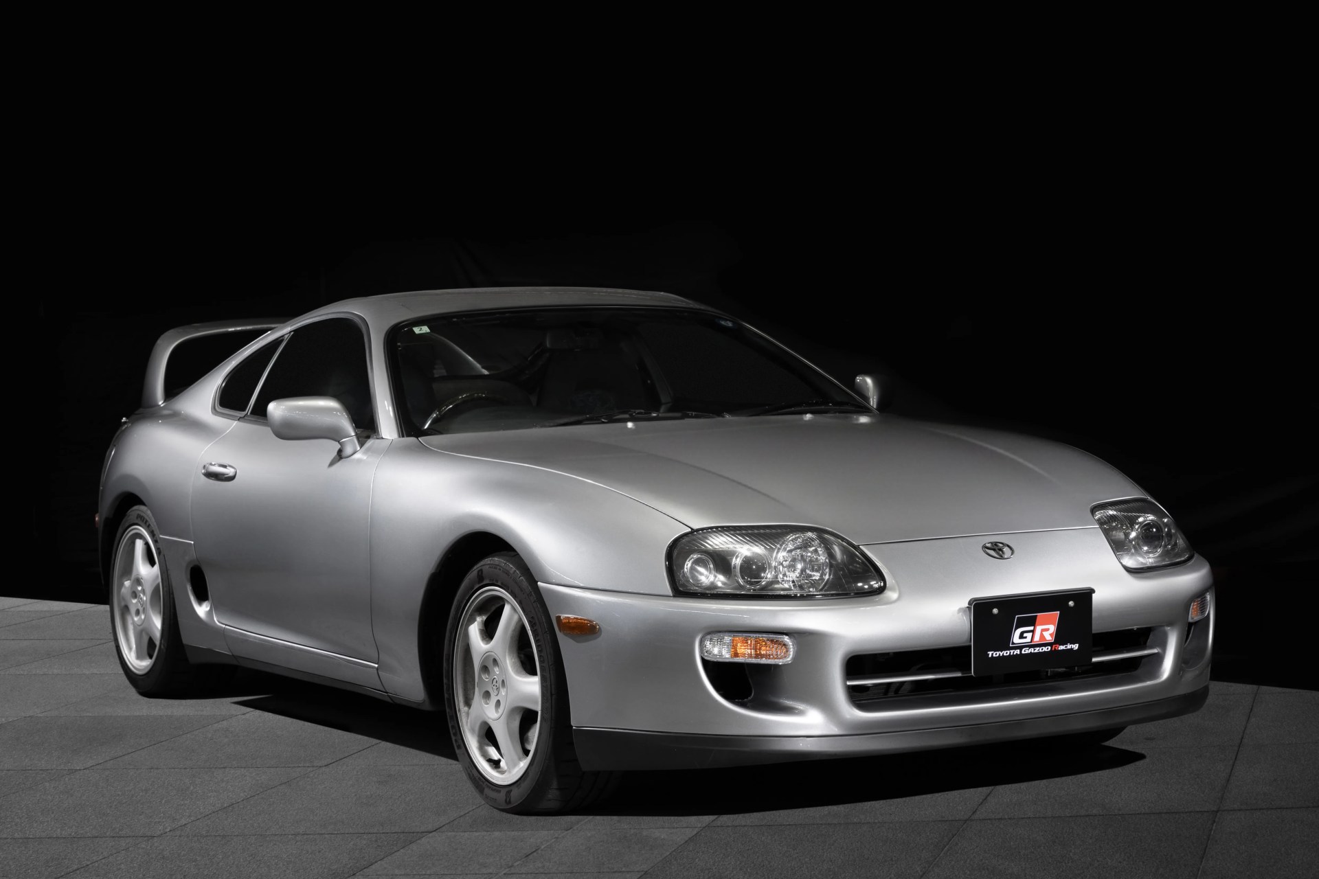 Silver Toyota Supra sports car with a rear spoiler and Toyota Gazoo Racing license plate on a dark background.