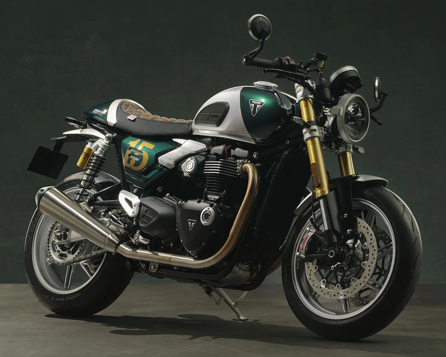 Green and silver Triumph motorcycle with gold front forks and a patterned seat on a gray floor.