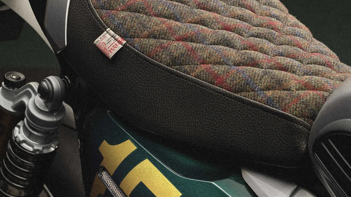 Quilted tweed motorcycle seat with a small fabric tag, mounted on a green bike frame with visible suspension.