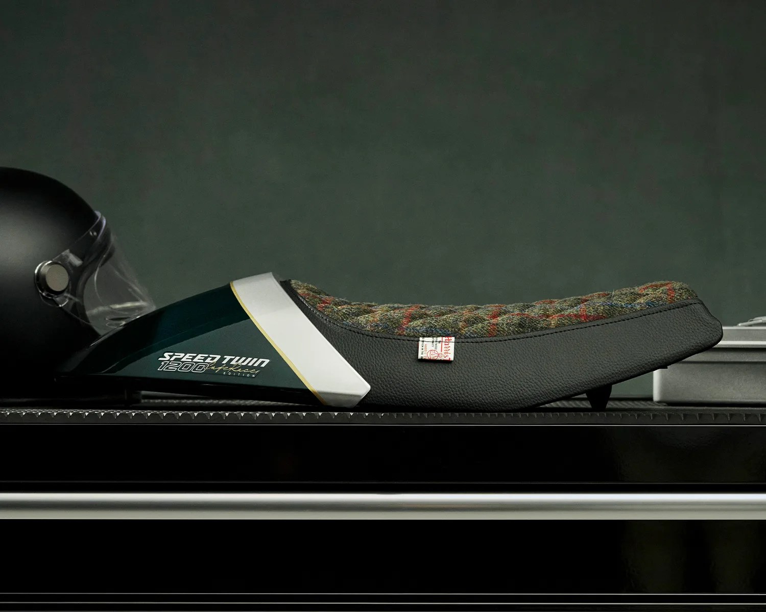 Motorcycle seat with black leather and multicolored tweed fabric, labeled "Speed Twin 1200" on a dark green panel.