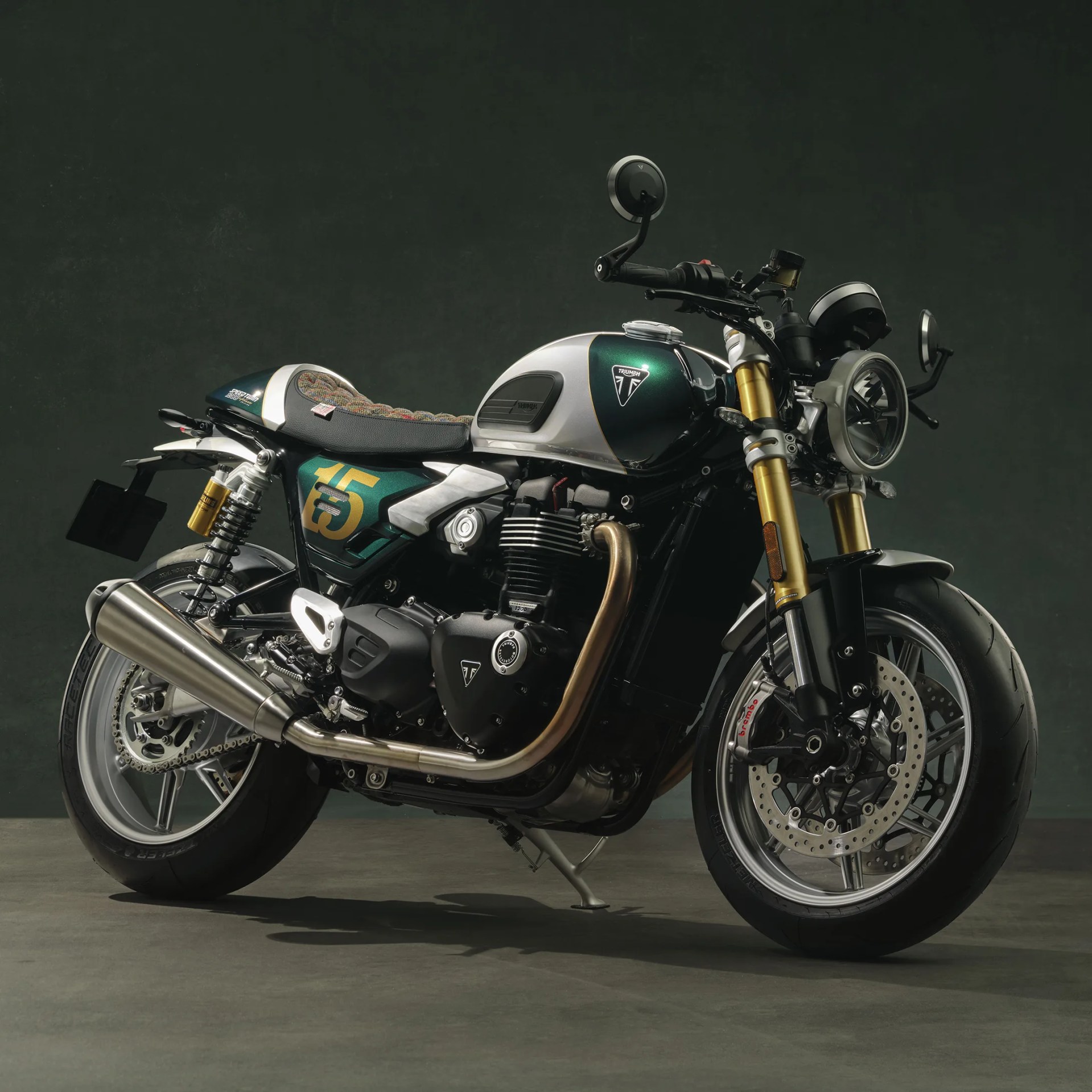 Green and silver Triumph motorcycle with gold front forks and a brown exhaust pipe on a gray floor.