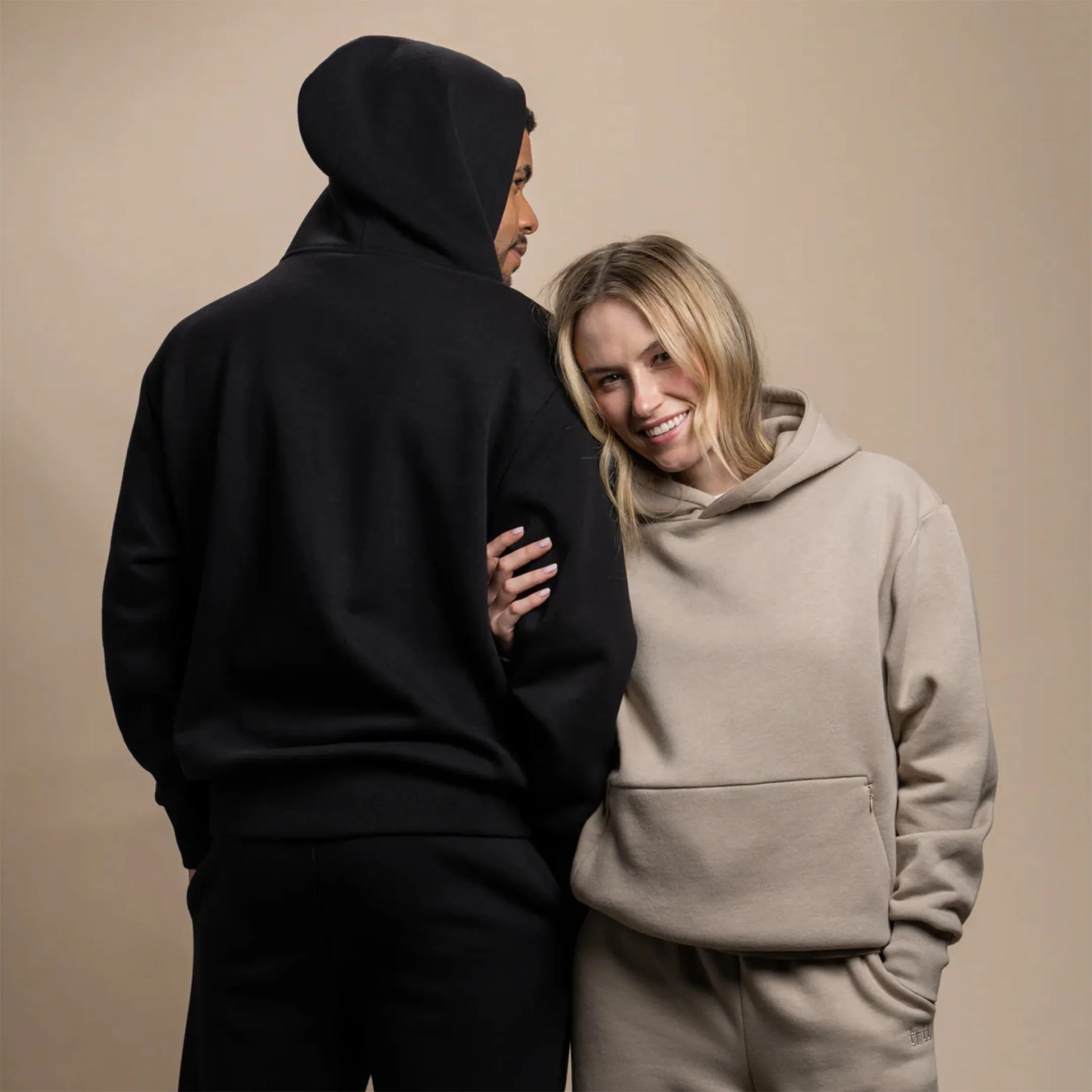 Man in a black hoodie and pants standing next to a smiling woman in a beige hoodie and matching pants.