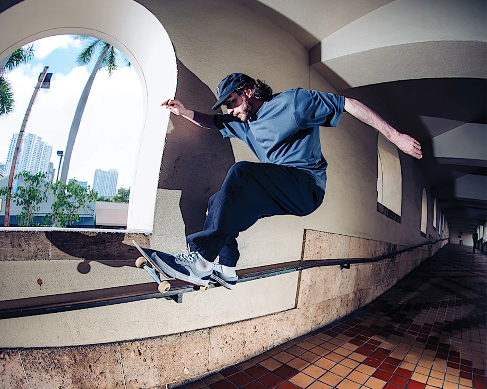 Skateboarder in blue shirt and cap grinding a metal rail in a tiled corridor with arched windows.