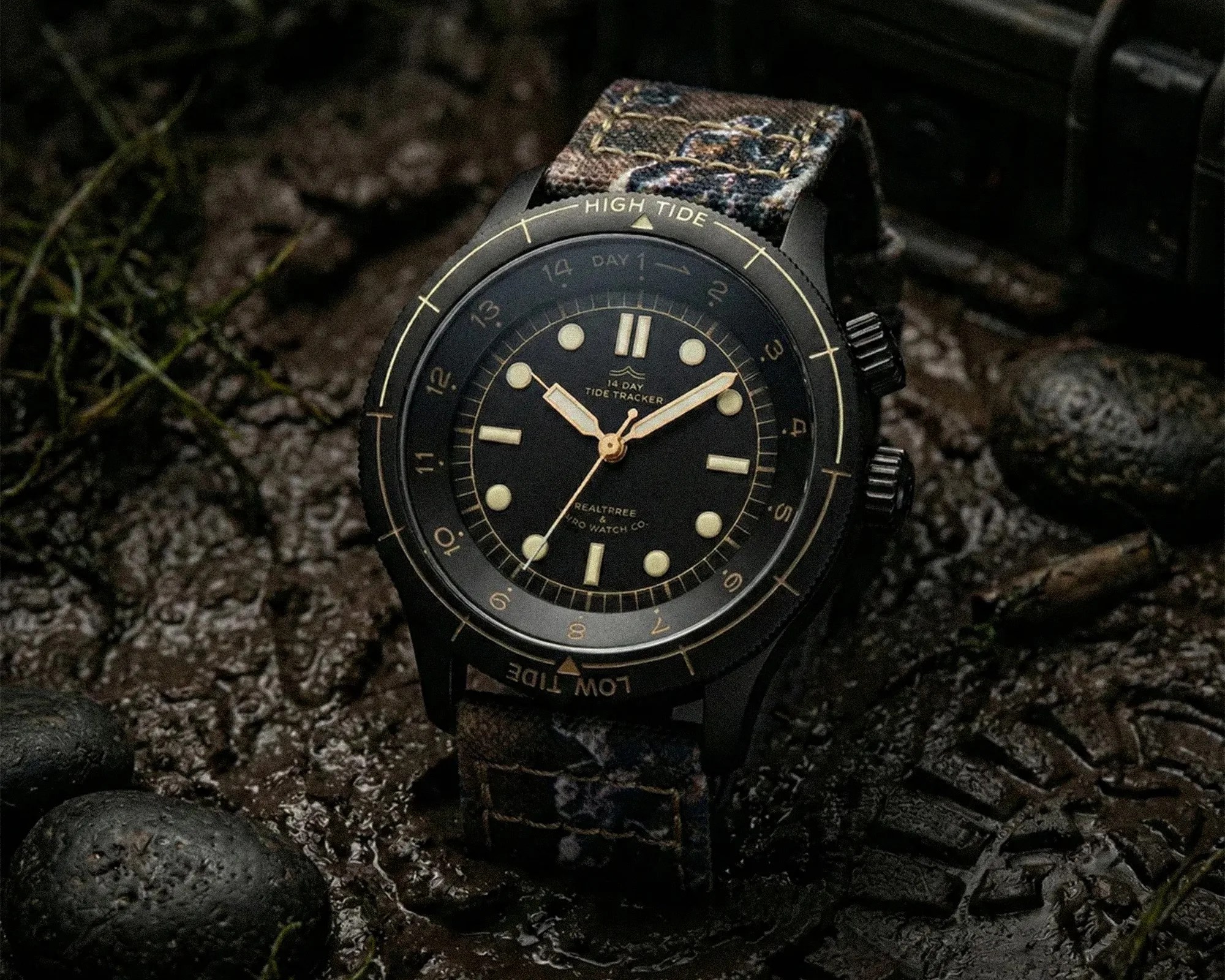 Black wristwatch with luminous markers and hands, fabric strap with camouflage pattern, placed on wet rocky ground.