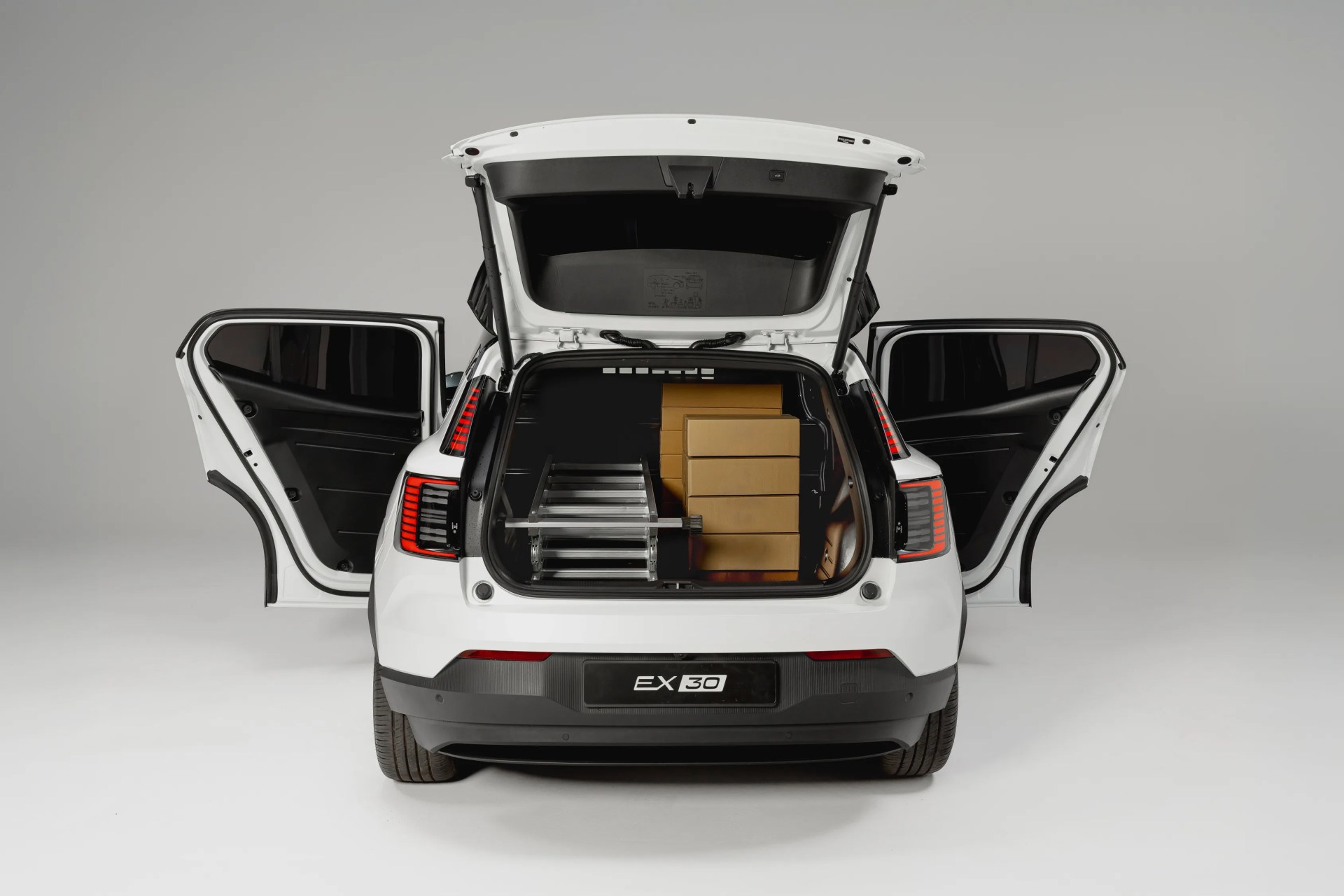 White EX30 car with rear hatch and side doors open, showing stacked cardboard boxes and a folded ladder in the trunk.