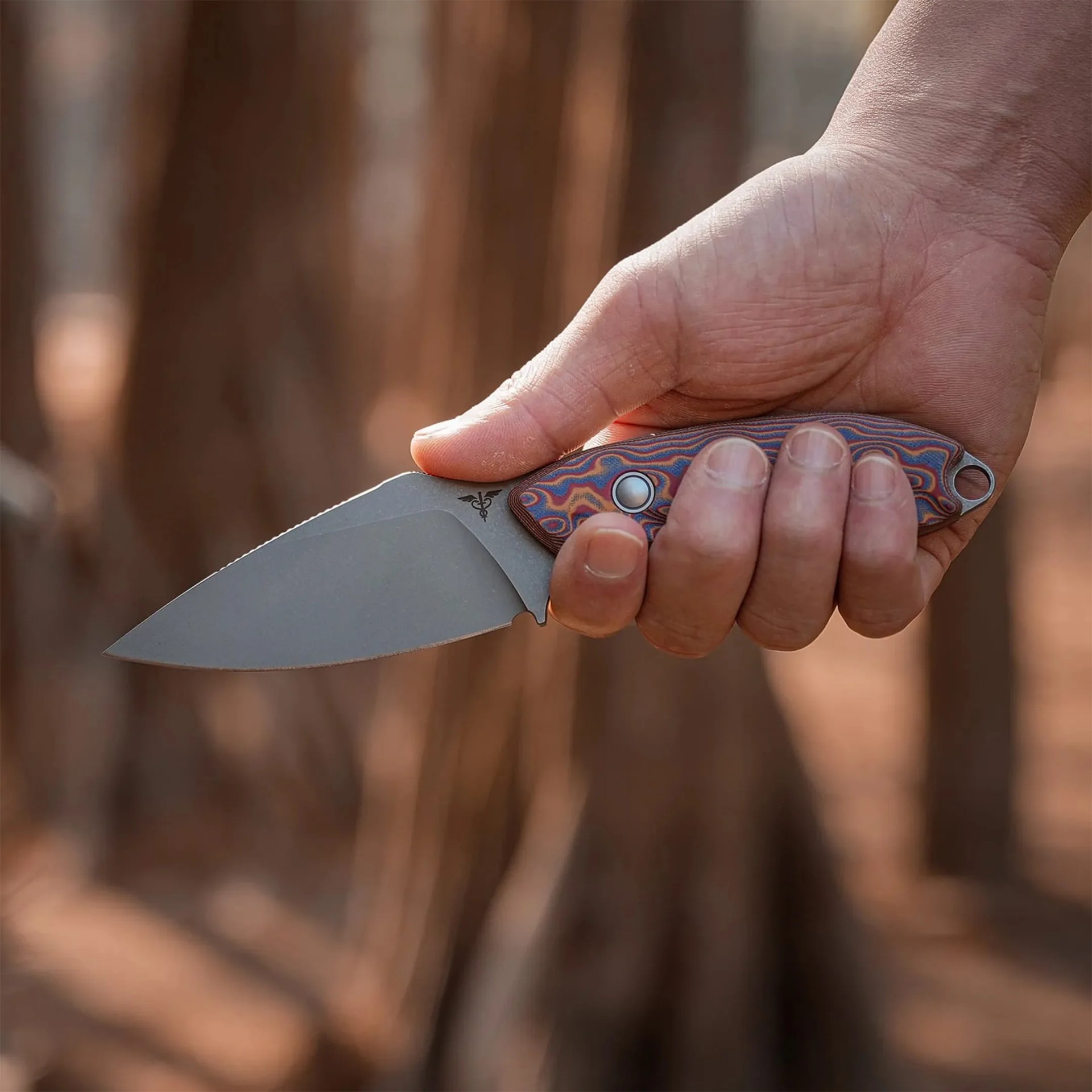 Hand holding a folding knife with a stonewashed blade and a colorful patterned handle.