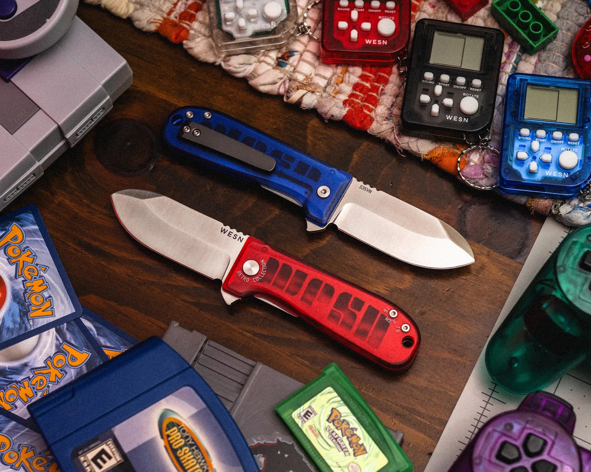 Two folding knives with silver blades and translucent handles, one blue and one red, placed on a wooden surface surrounded by retro gaming items.