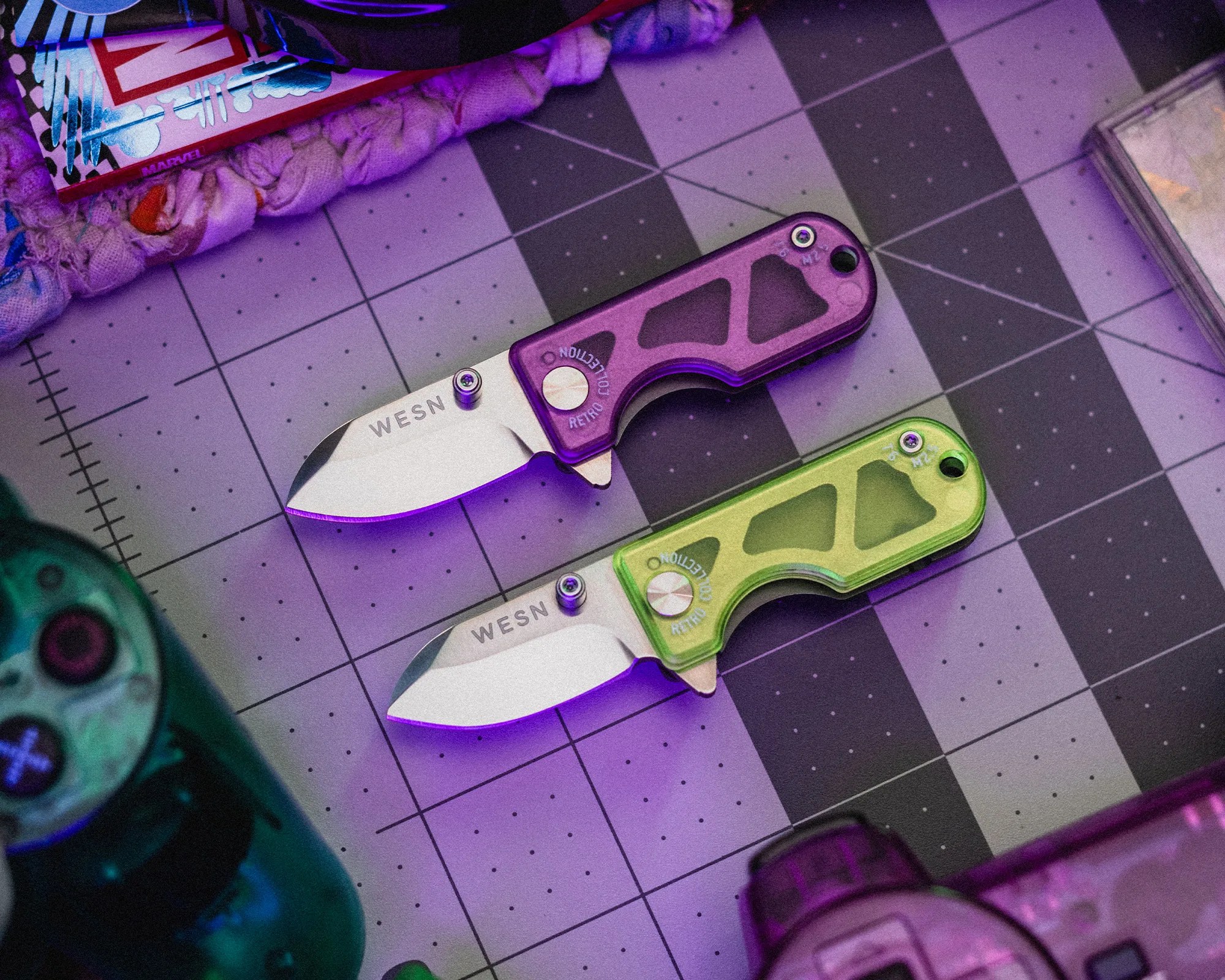 Two small folding knives with stainless steel blades and translucent handles, one purple and one green, on a cutting mat.