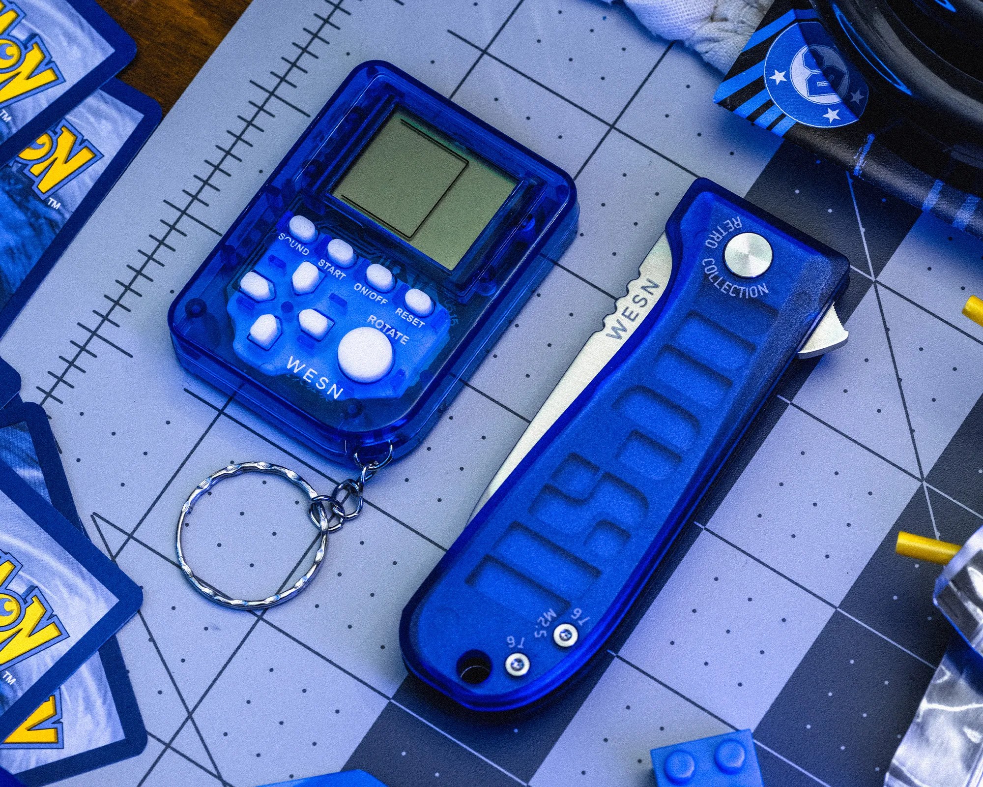 Blue translucent handheld electronic game with keychain and blue translucent folding knife on a cutting mat.