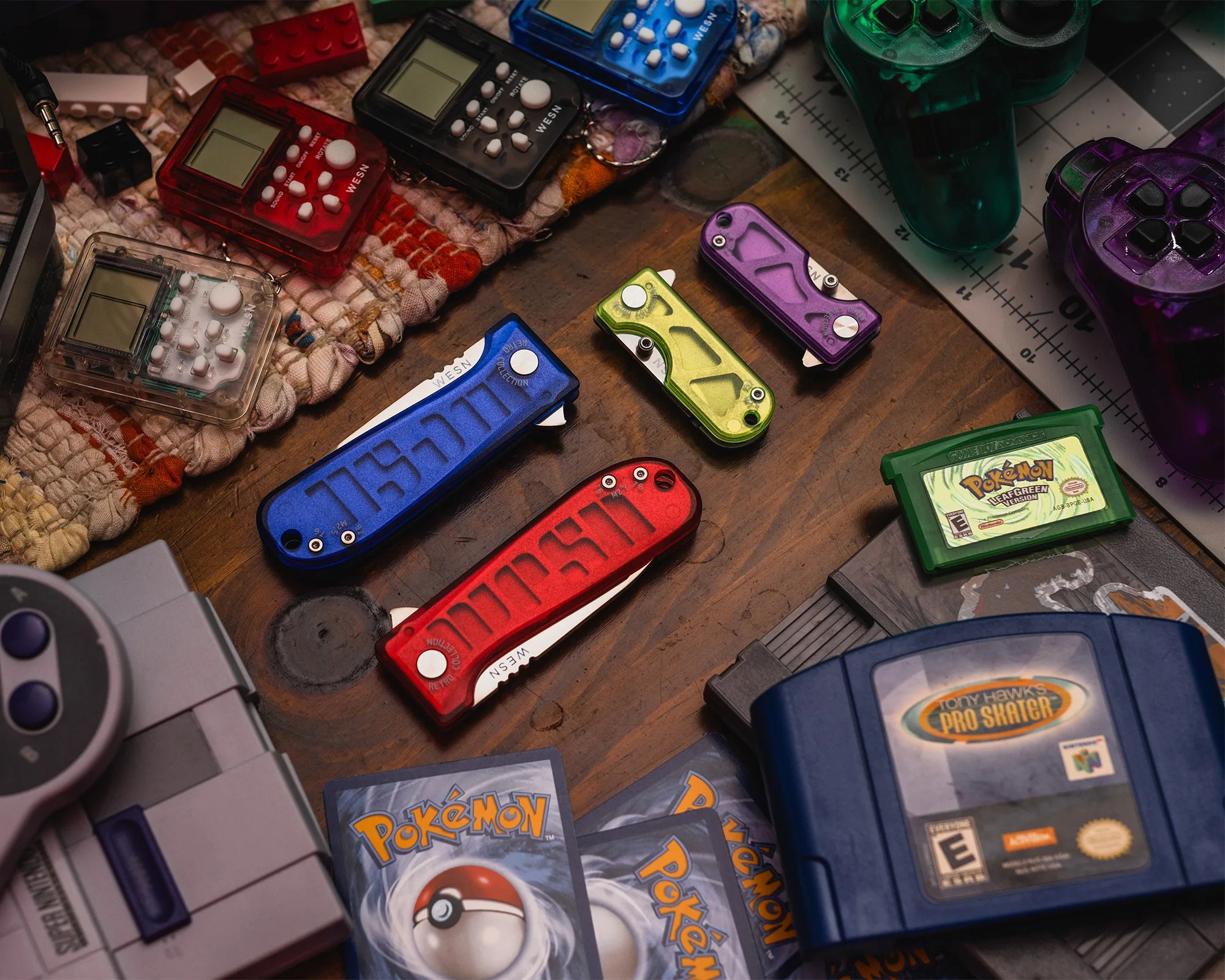 Collection of colorful WESN folding knives, retro handheld game consoles, Pok&eacute;mon cards, and game cartridges on a wooden surface.