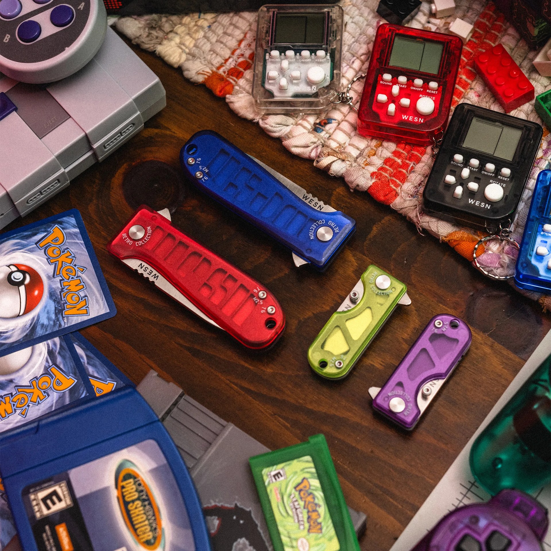 Collection of colorful retro-style pocket knives and handheld electronic games on a wooden surface with Pok&eacute;mon cards and game cartridges.