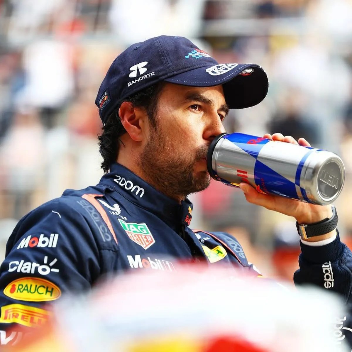 Person in a dark blue racing suit and cap drinking from a silver and blue Red Bull can.