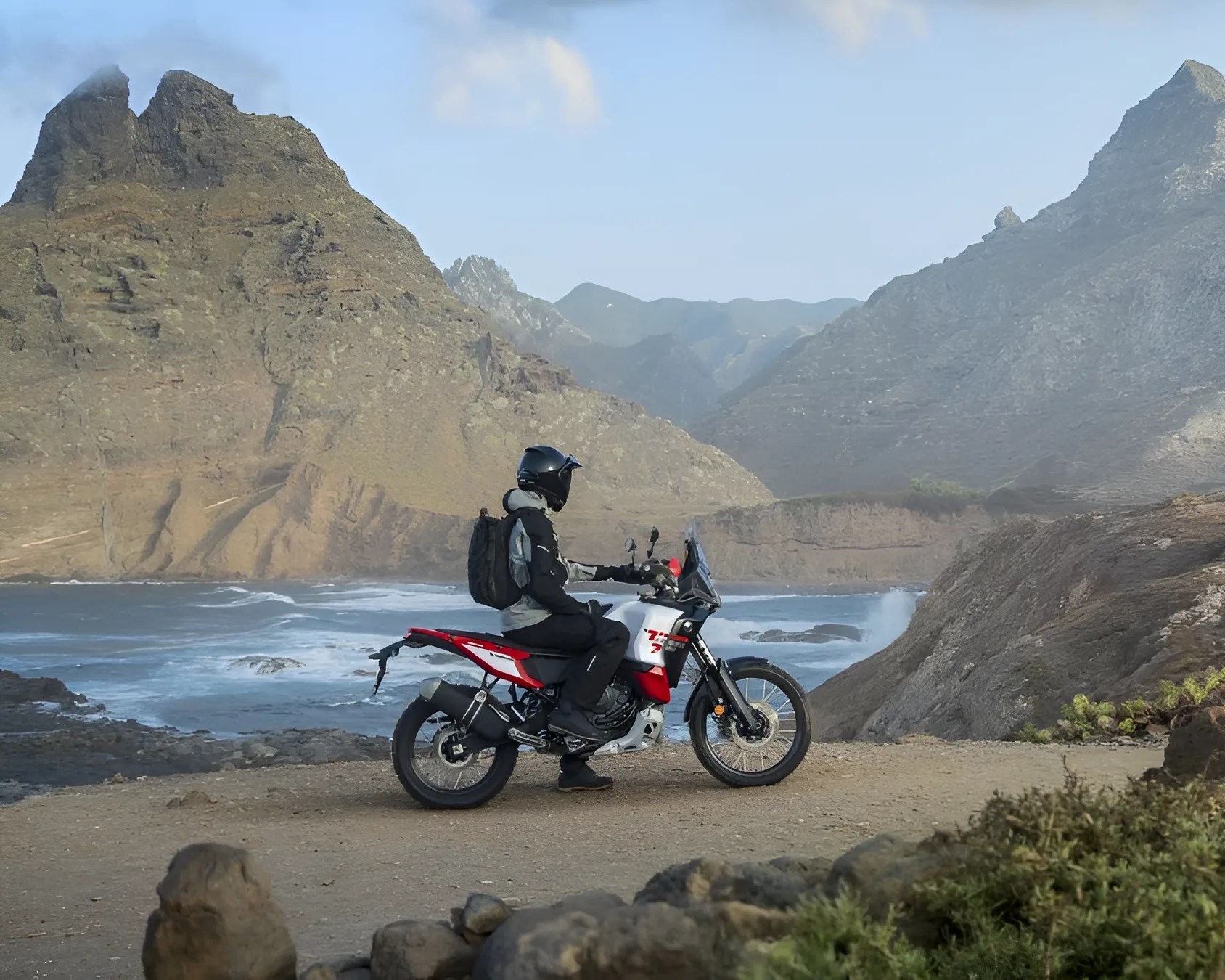 Person wearing black and gray riding gear and helmet sitting on a red and white adventure motorcycle near rocky coastline with mountains in the background.