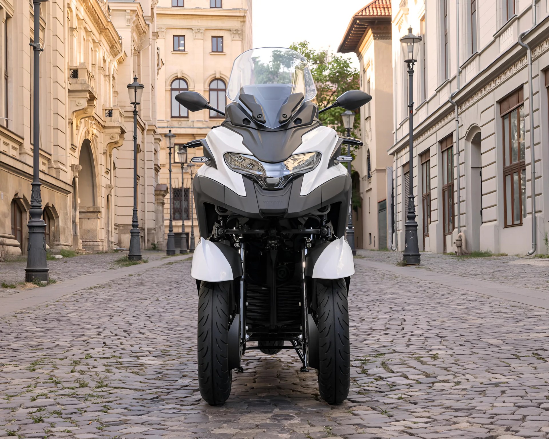 White and black three-wheeled motorcycle with a windshield parked on a cobblestone street between historic buildings.