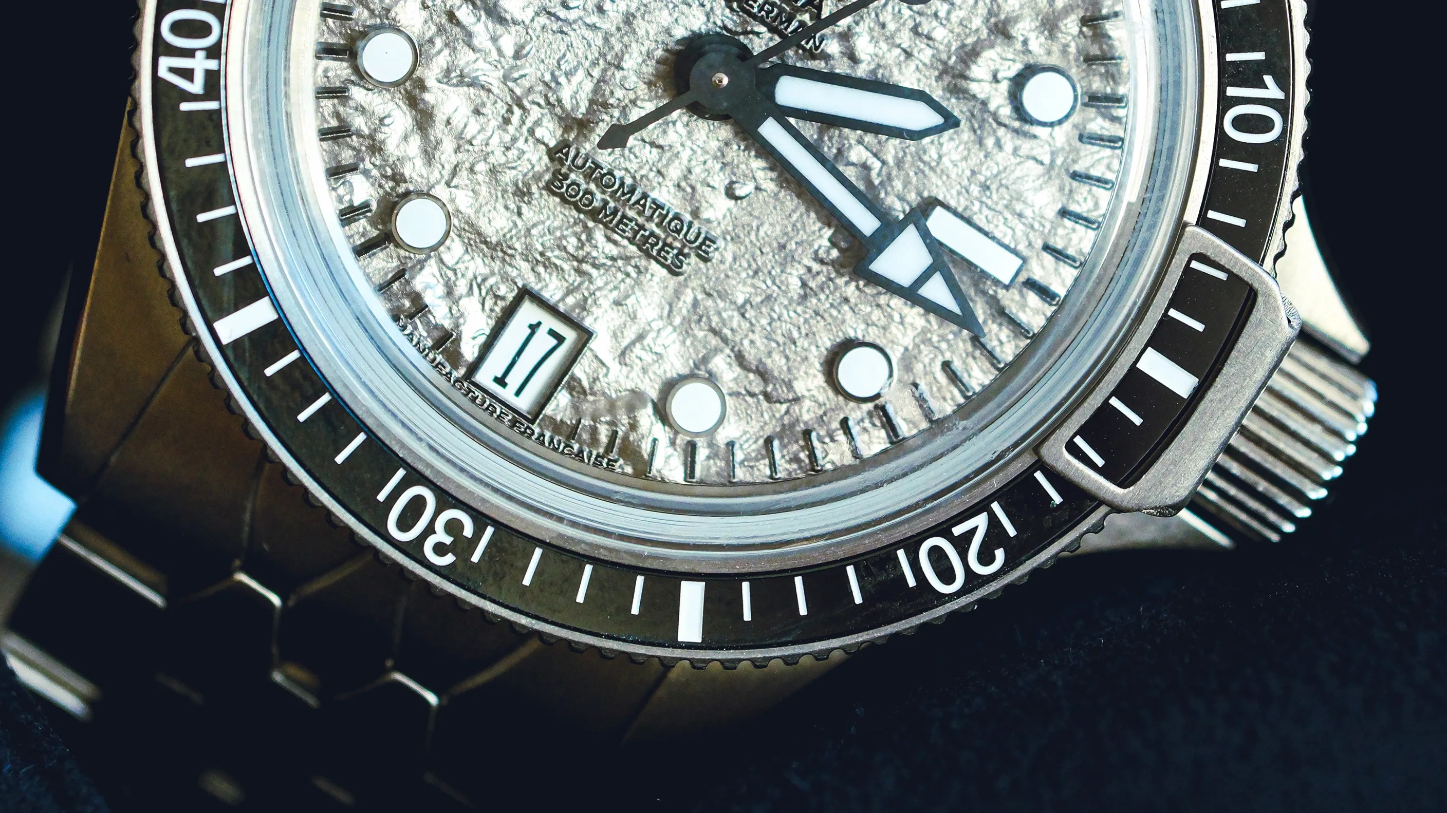 Close-up of a silver textured dial watch with black and white bezel, luminous hands, date window showing 17, and metal bracelet.