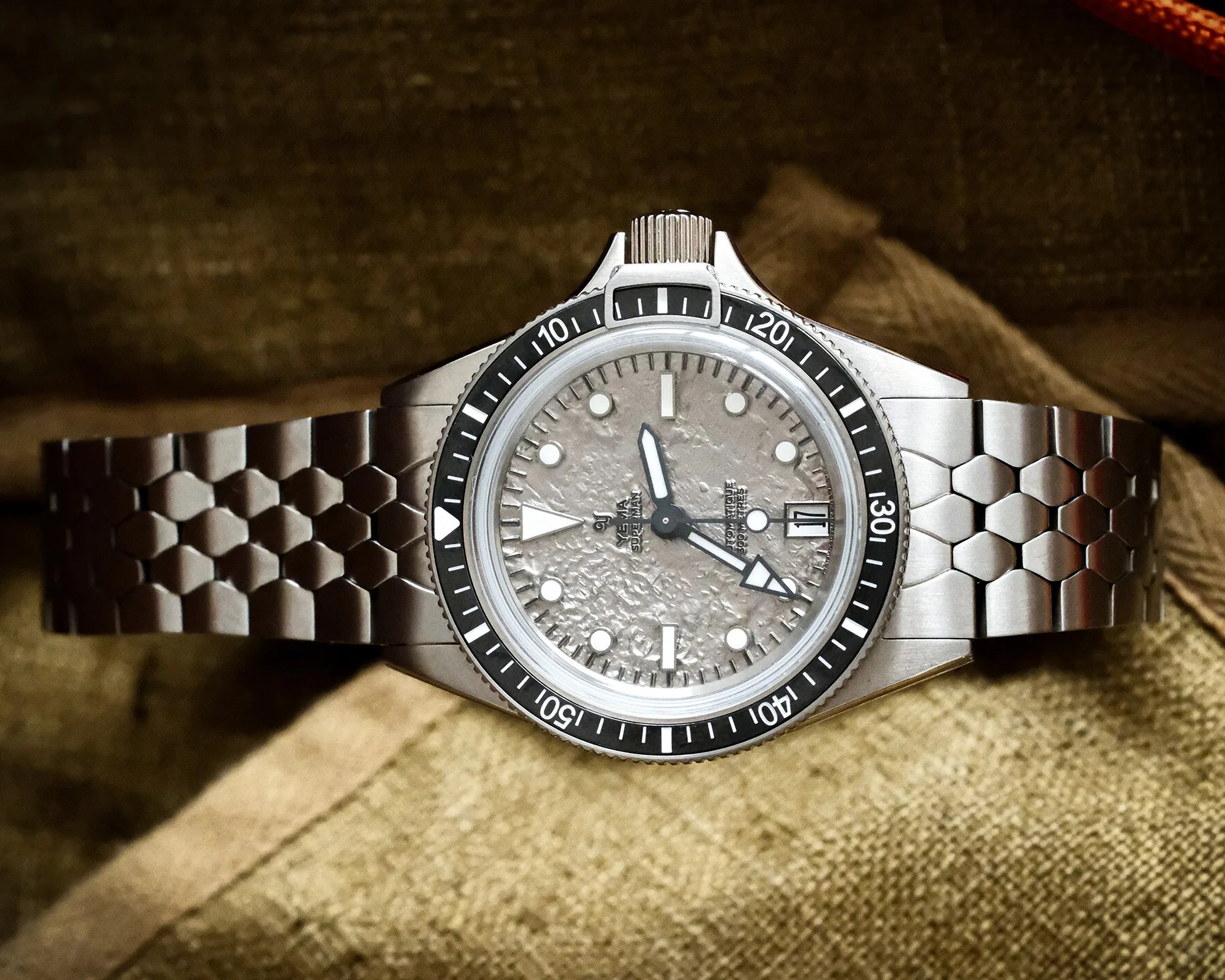 Stainless steel dive watch with textured silver dial, black bezel, and hexagonal-link bracelet on a brown fabric background.