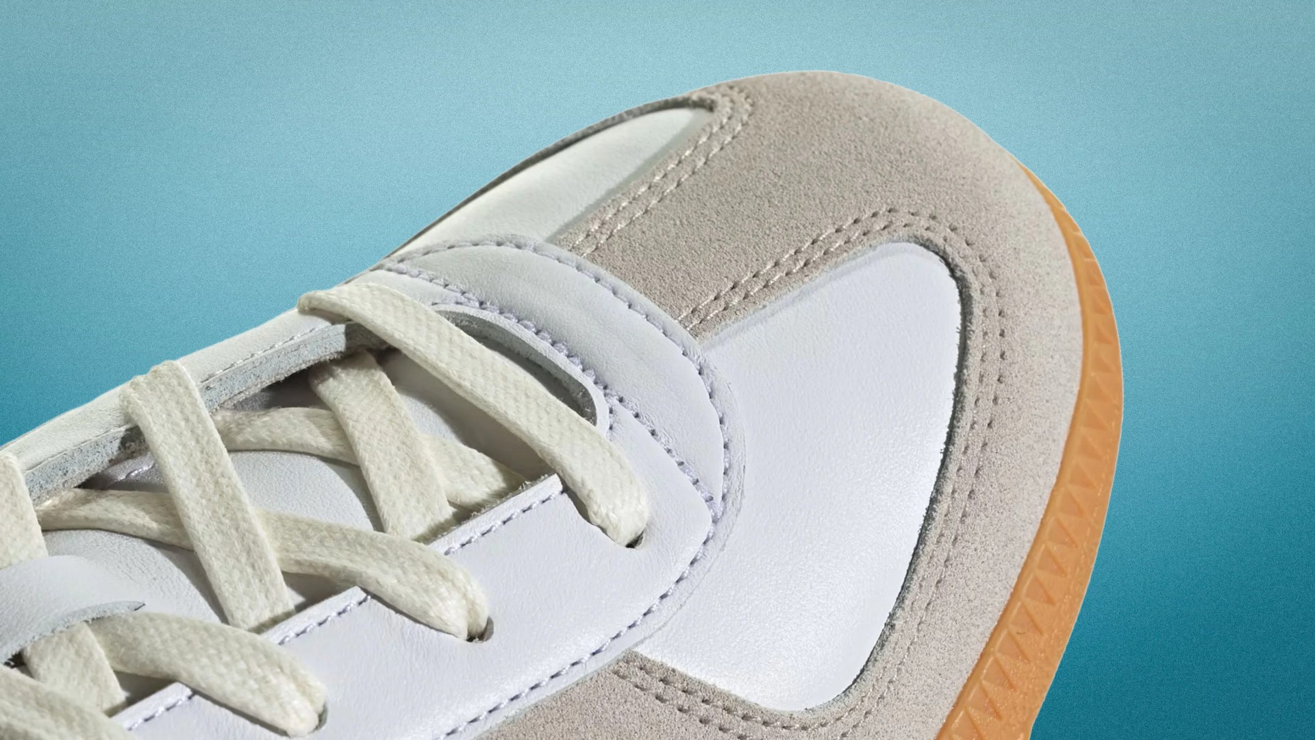 Close-up of a white and beige suede sneaker toe with cream laces and a gum rubber sole on a blue gradient background.