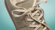 Close-up of a beige sneaker with cream laces, red stitching detail, and a gum sole against a teal background.