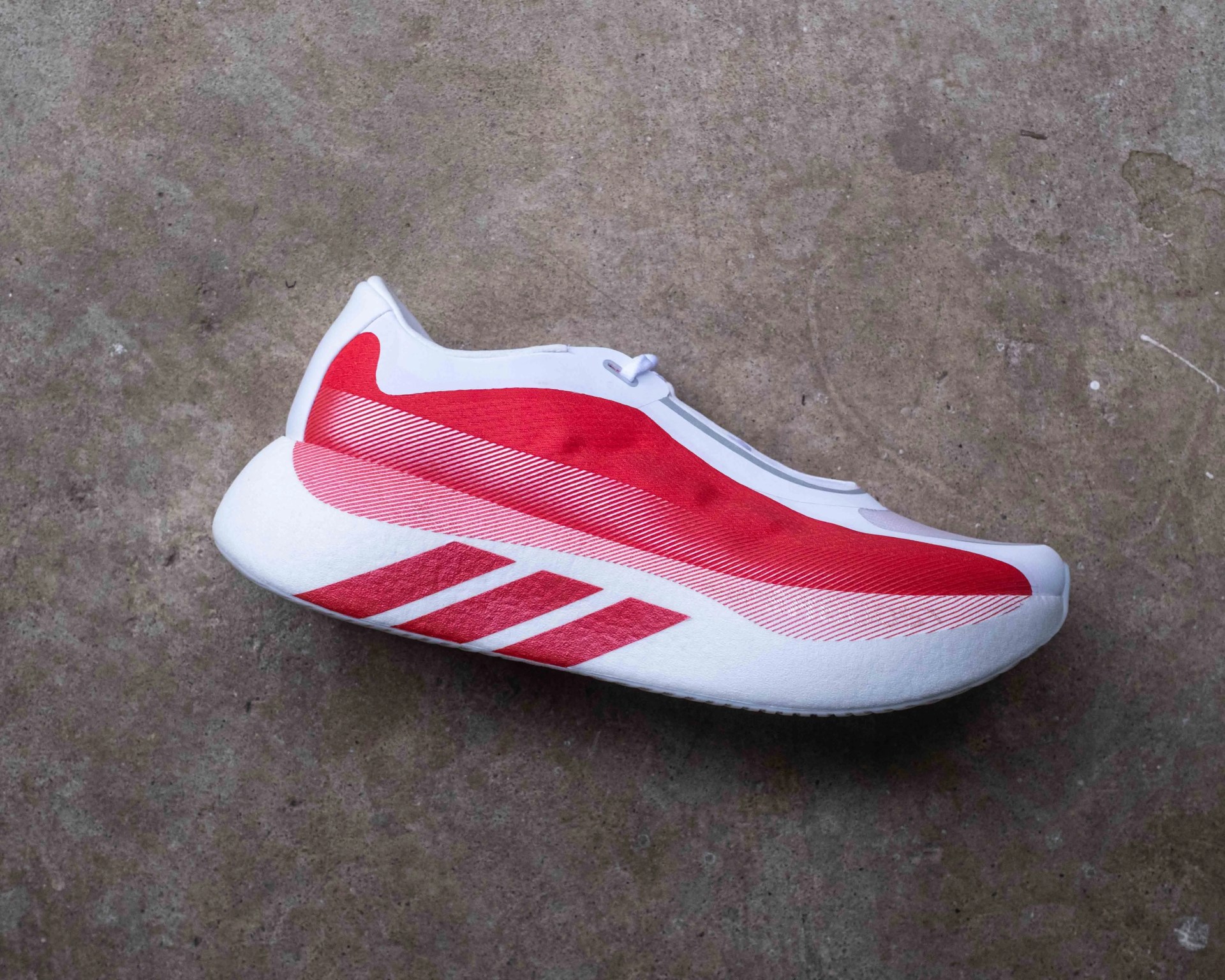 White and red athletic shoe with a thick sole and three red stripes on the side, placed on a concrete surface.