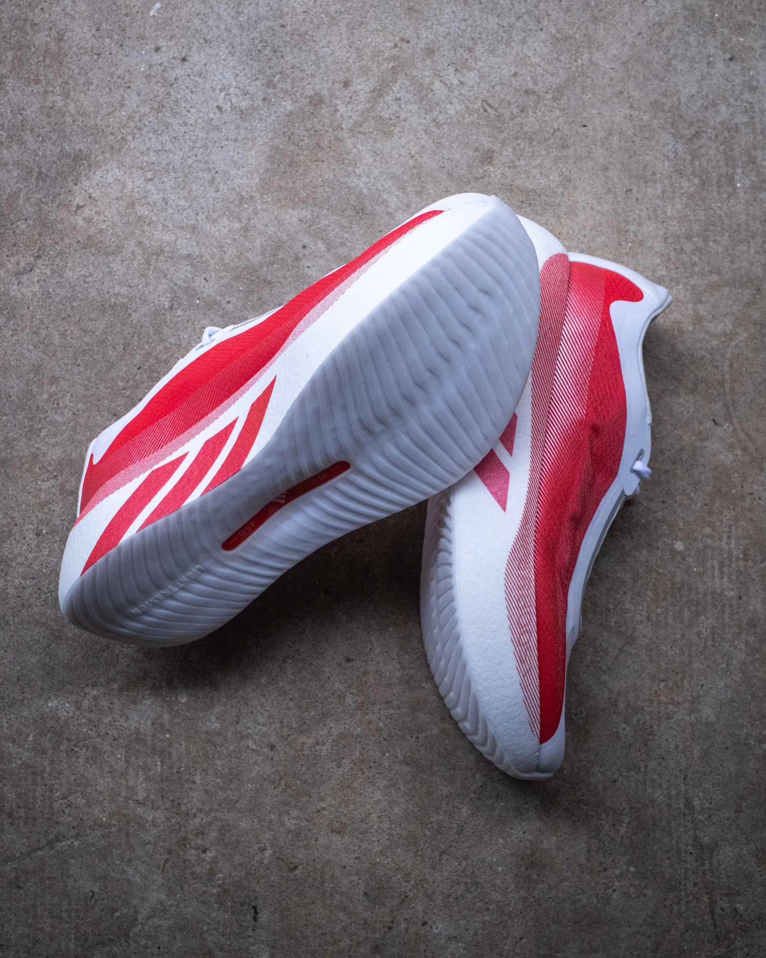 Pair of white and red athletic shoes with textured white soles on a concrete floor.