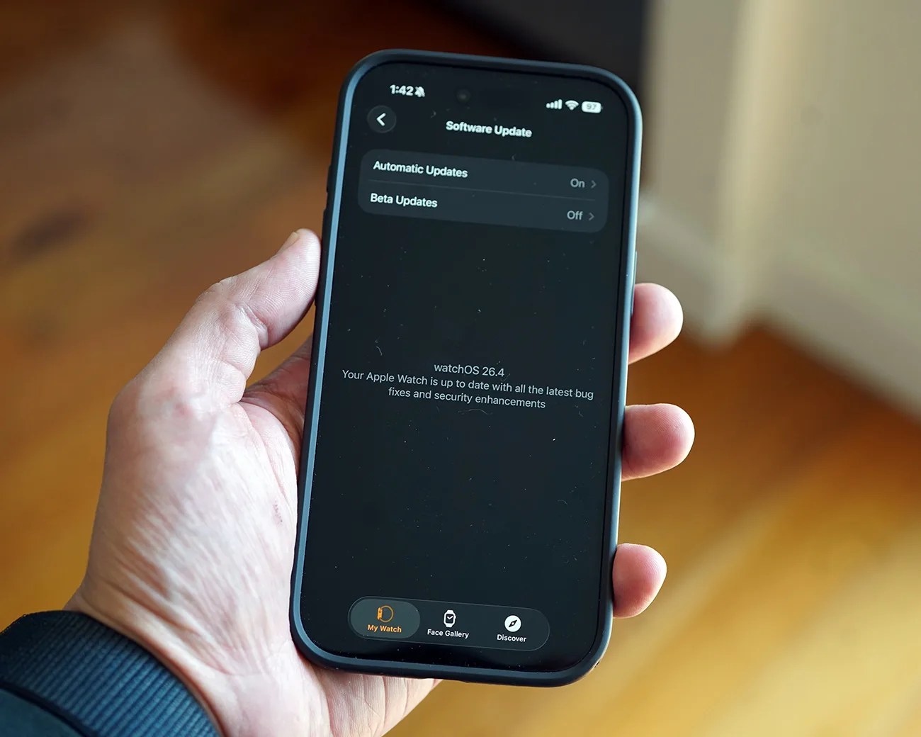 Hand holding a smartphone displaying a software update screen for watchOS 26.4 with automatic updates on.