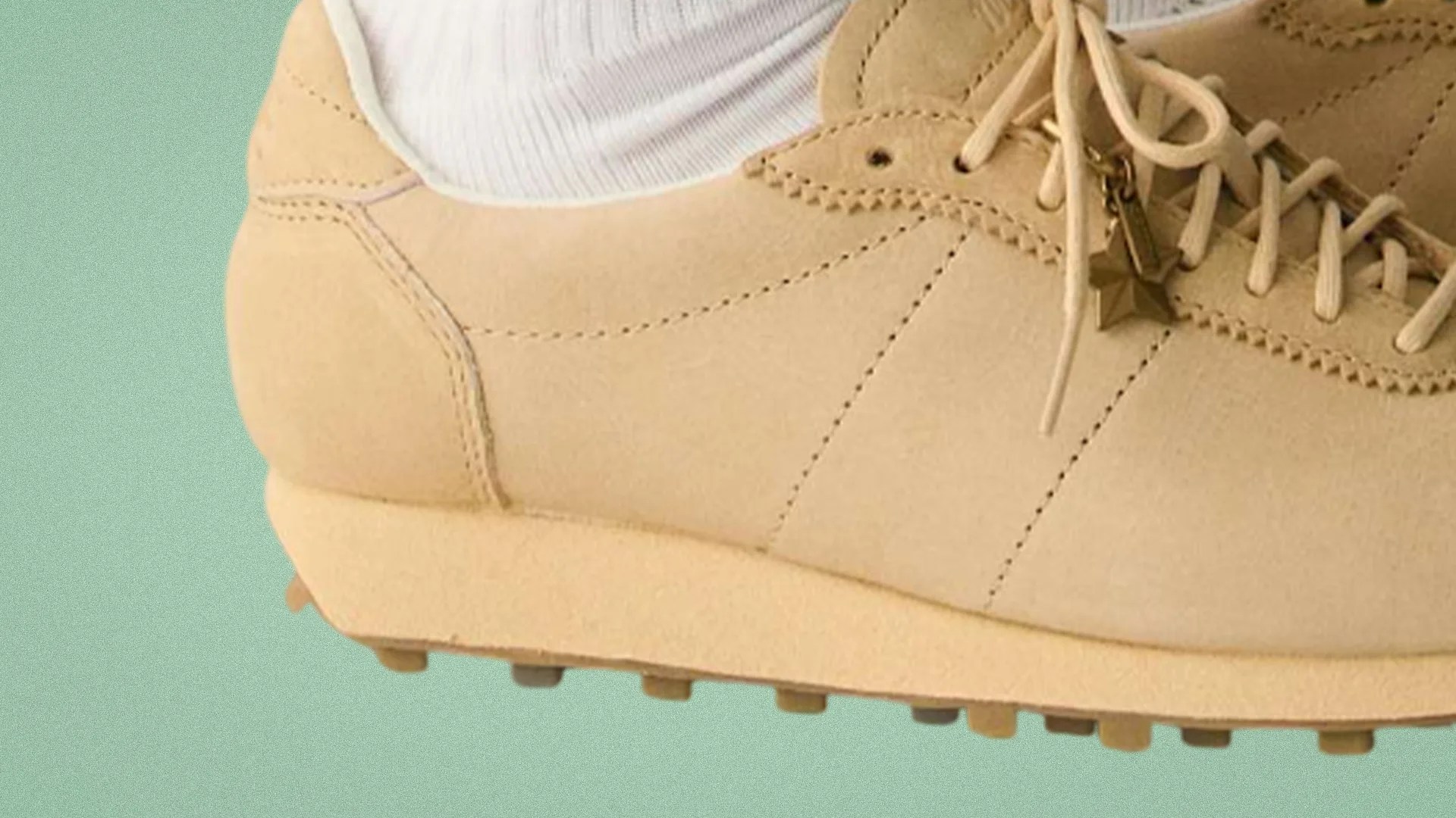 Beige suede sneaker with cream laces and star-shaped zipper charm worn with white socks.