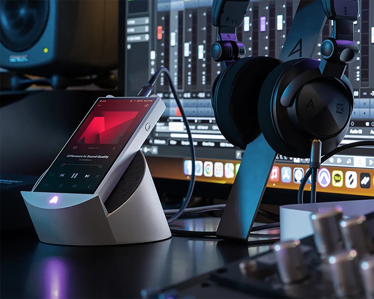 Silver digital audio player on a gray angled dock with black headphones hanging on a stand beside it.