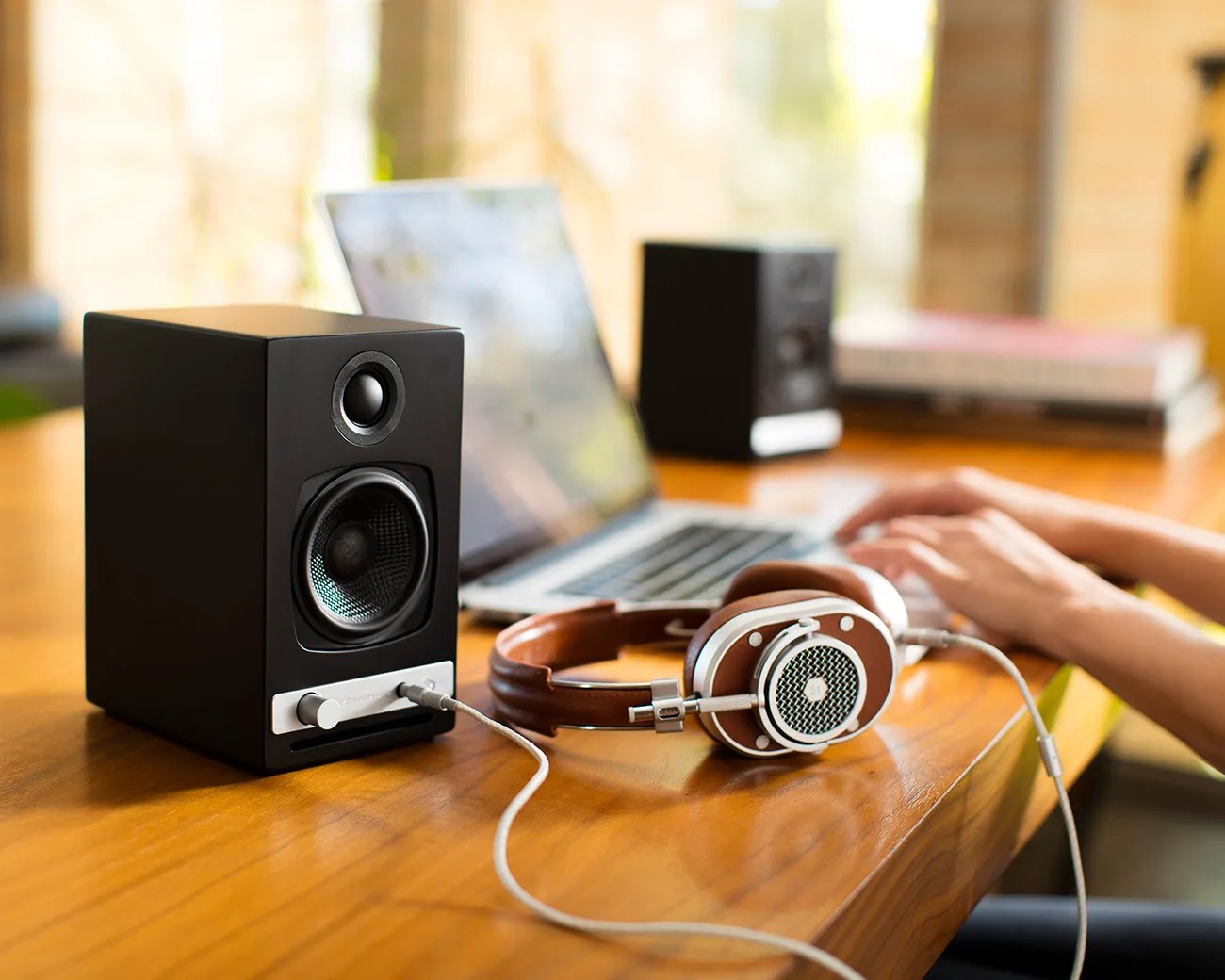 Black bookshelf speaker with silver control knob and connected brown and silver headphones on a wooden desk with a laptop in the background.