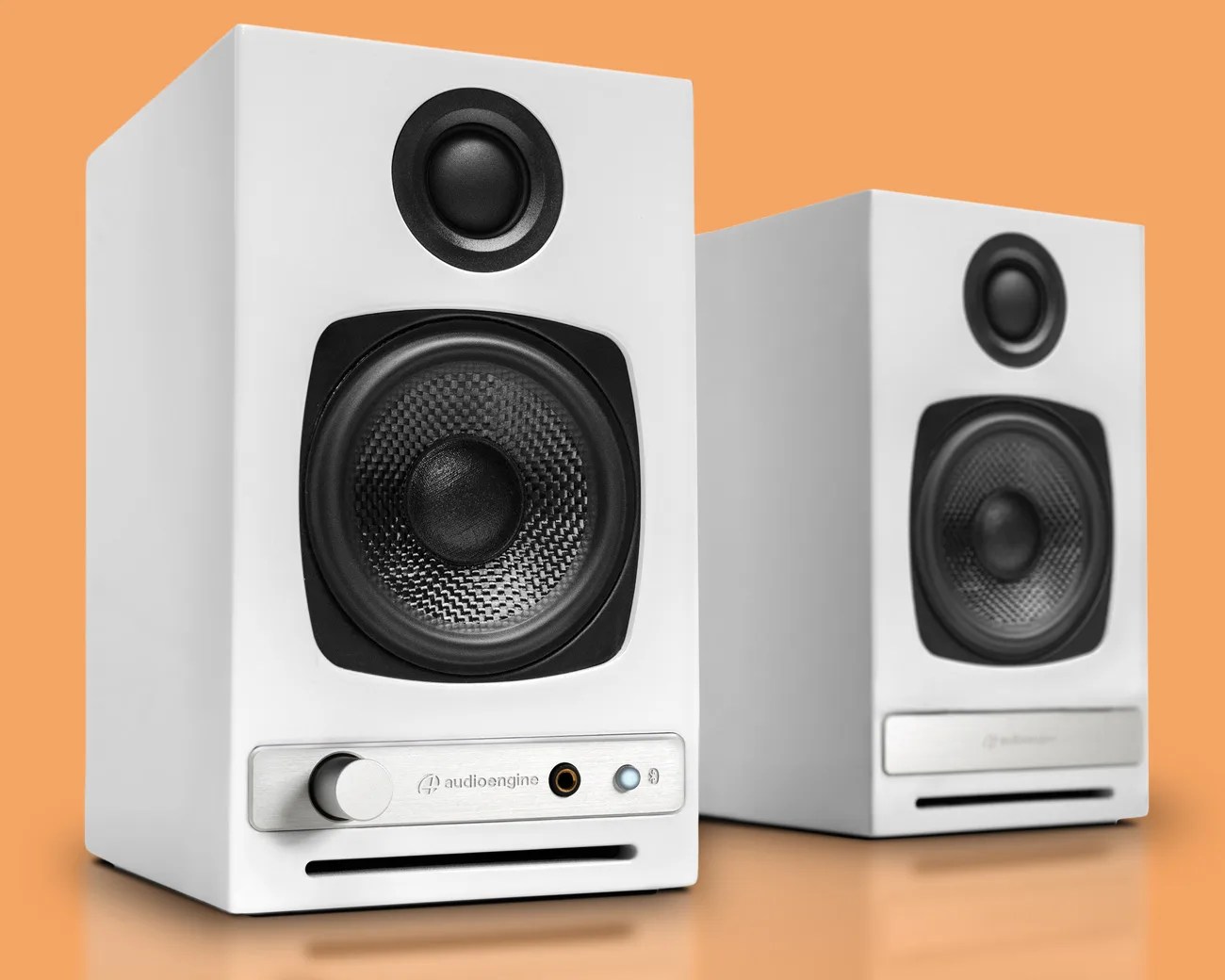 Pair of white Audioengine speakers with black drivers and silver control panel on an orange surface.