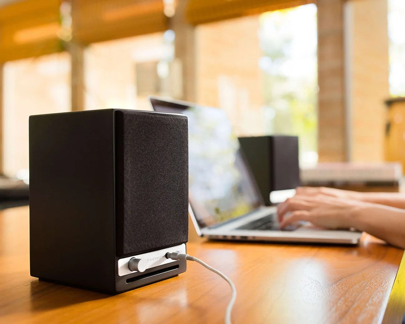 Black desktop speaker with fabric front and silver control knobs connected to a laptop on a wooden desk.