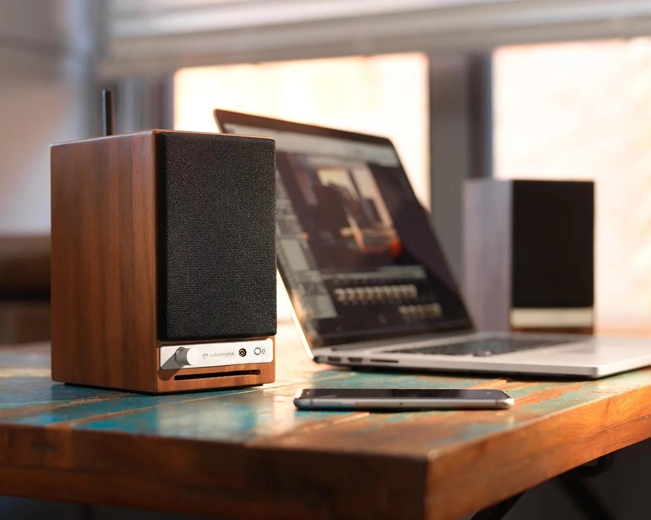 Wooden Audioengine speaker with black grille and silver control panel on a rustic wooden table next to a laptop and smartphone.