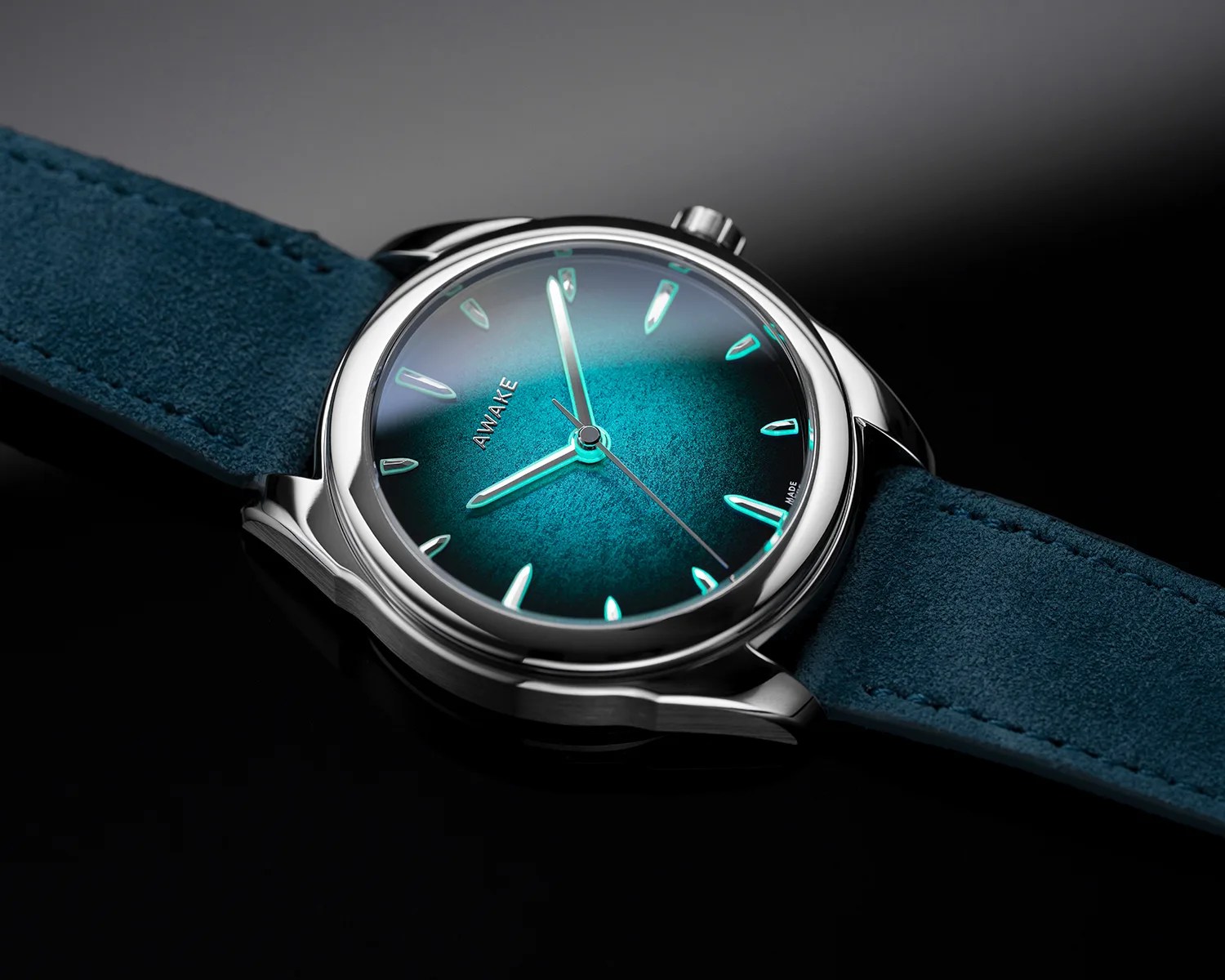 Silver wristwatch with a textured teal dial, luminous hands and markers, and a teal suede strap on a black surface.