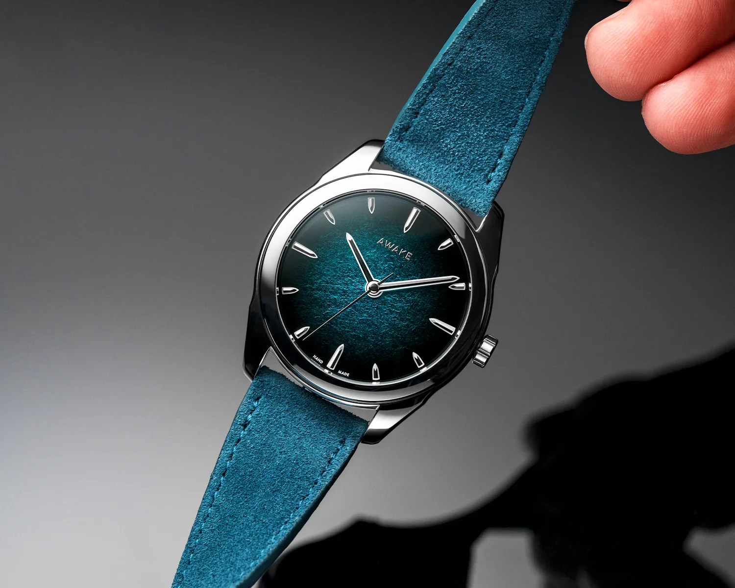 Silver wristwatch with a textured teal dial and matching suede strap held by a hand.