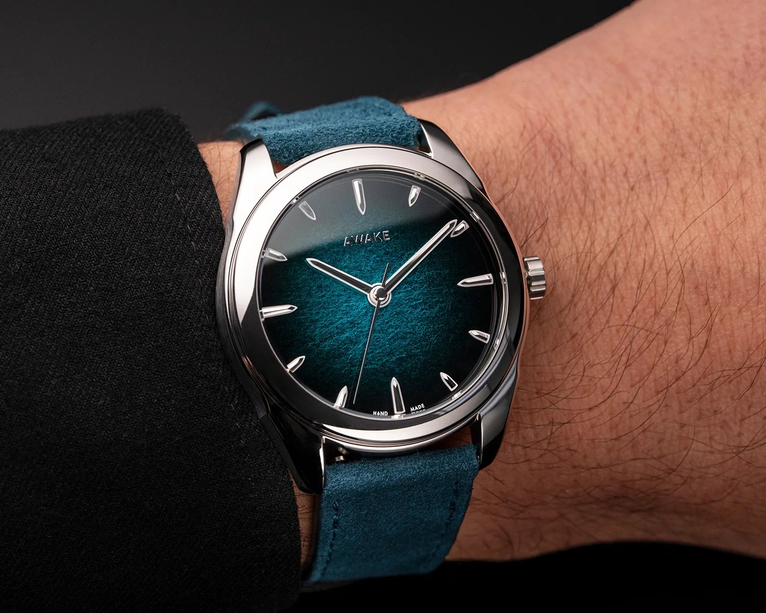 Silver wristwatch with a textured teal dial and matching suede strap worn on a wrist.