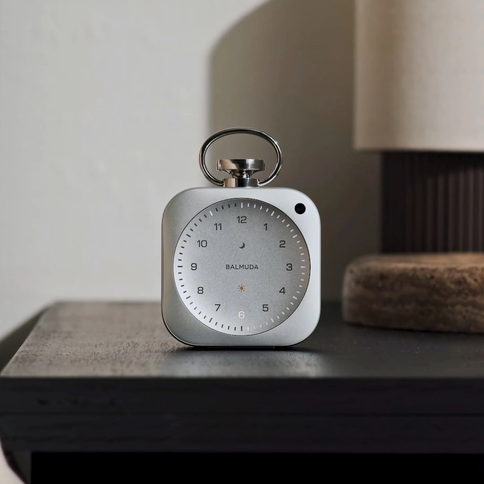 Silver Balmuda analog clock with rounded square shape on a dark wooden surface.