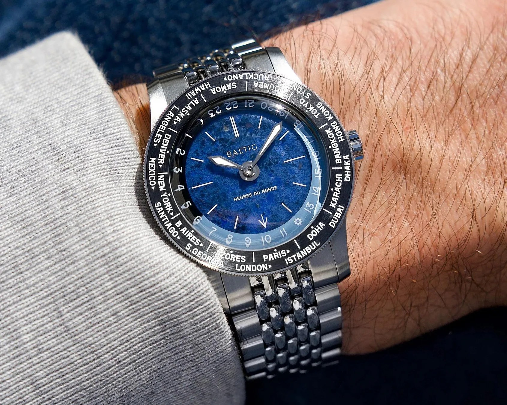 Silver metal wristwatch with a blue textured dial and world city names on the bezel, worn on a wrist.