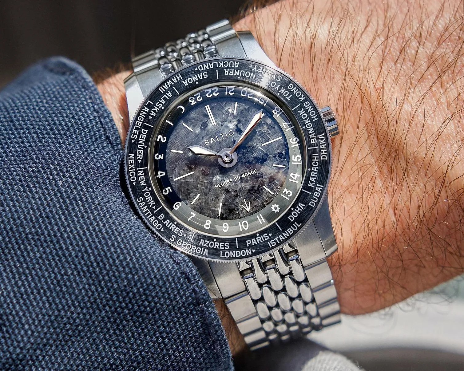 Silver Baltic wristwatch with a textured dark dial and world city names on the bezel, worn on a wrist.