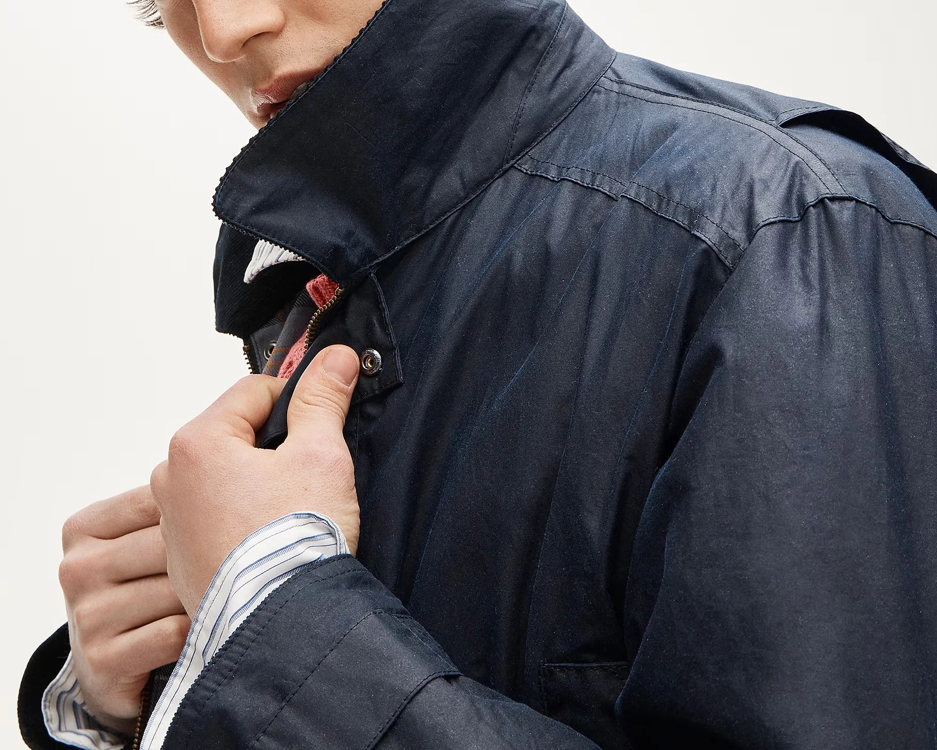 Close-up of a person fastening the snap button on a dark navy jacket with a high collar, wearing a striped shirt underneath.