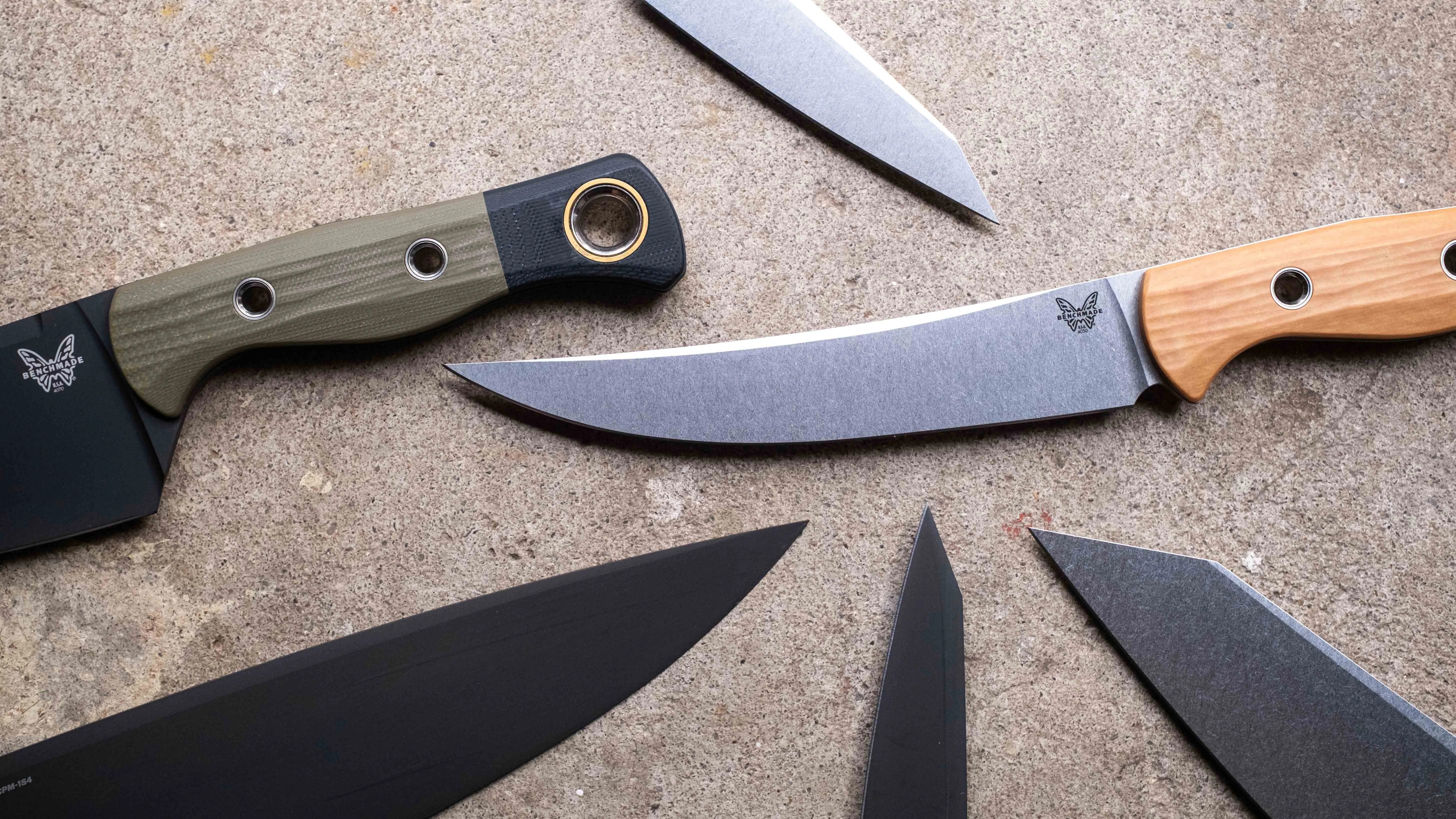 Five knives with various blade shapes and handle materials, including wood and textured synthetic, arranged on a stone surface.