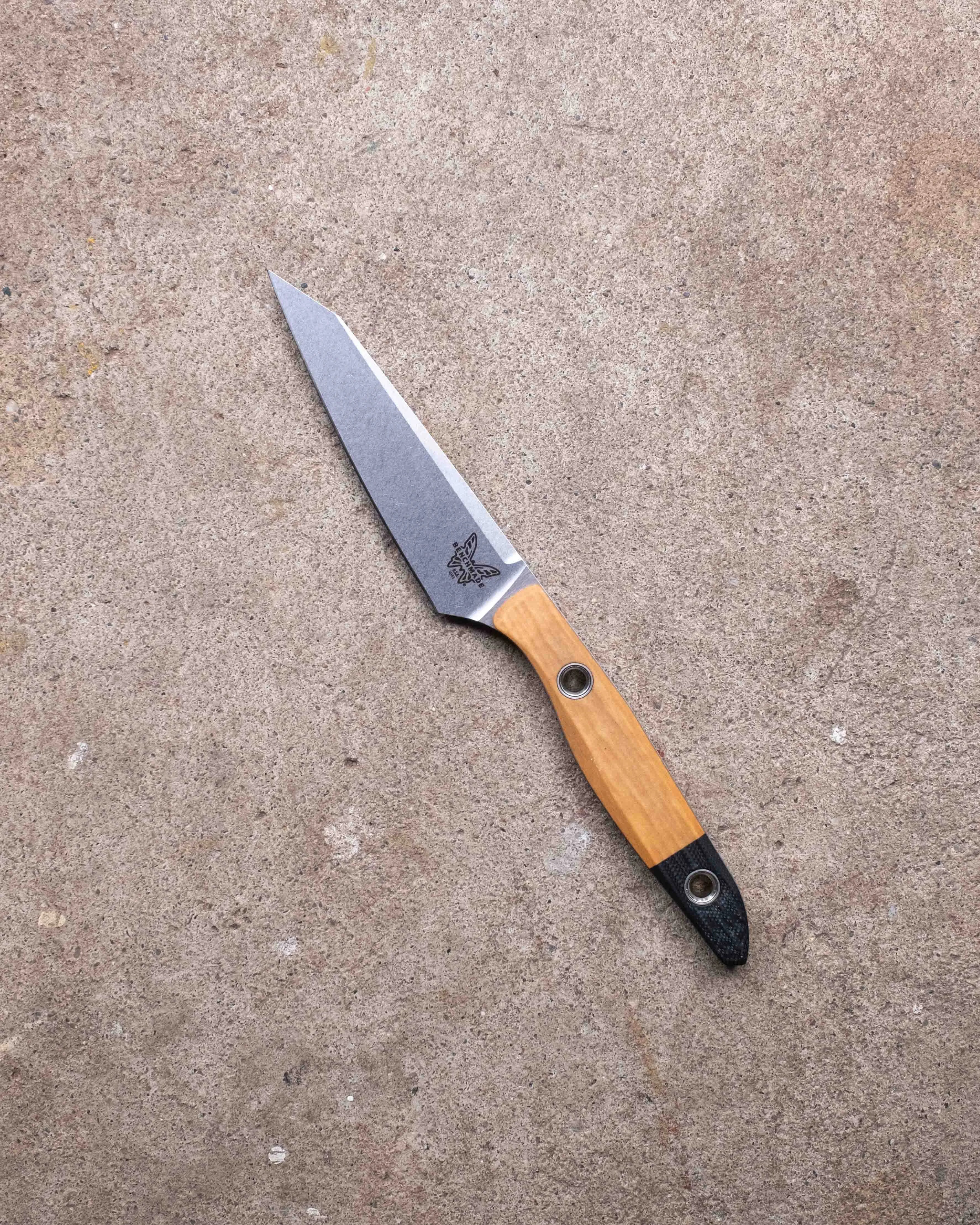 Knife with a stonewashed blade, wooden handle, and black textured end resting on a concrete surface.