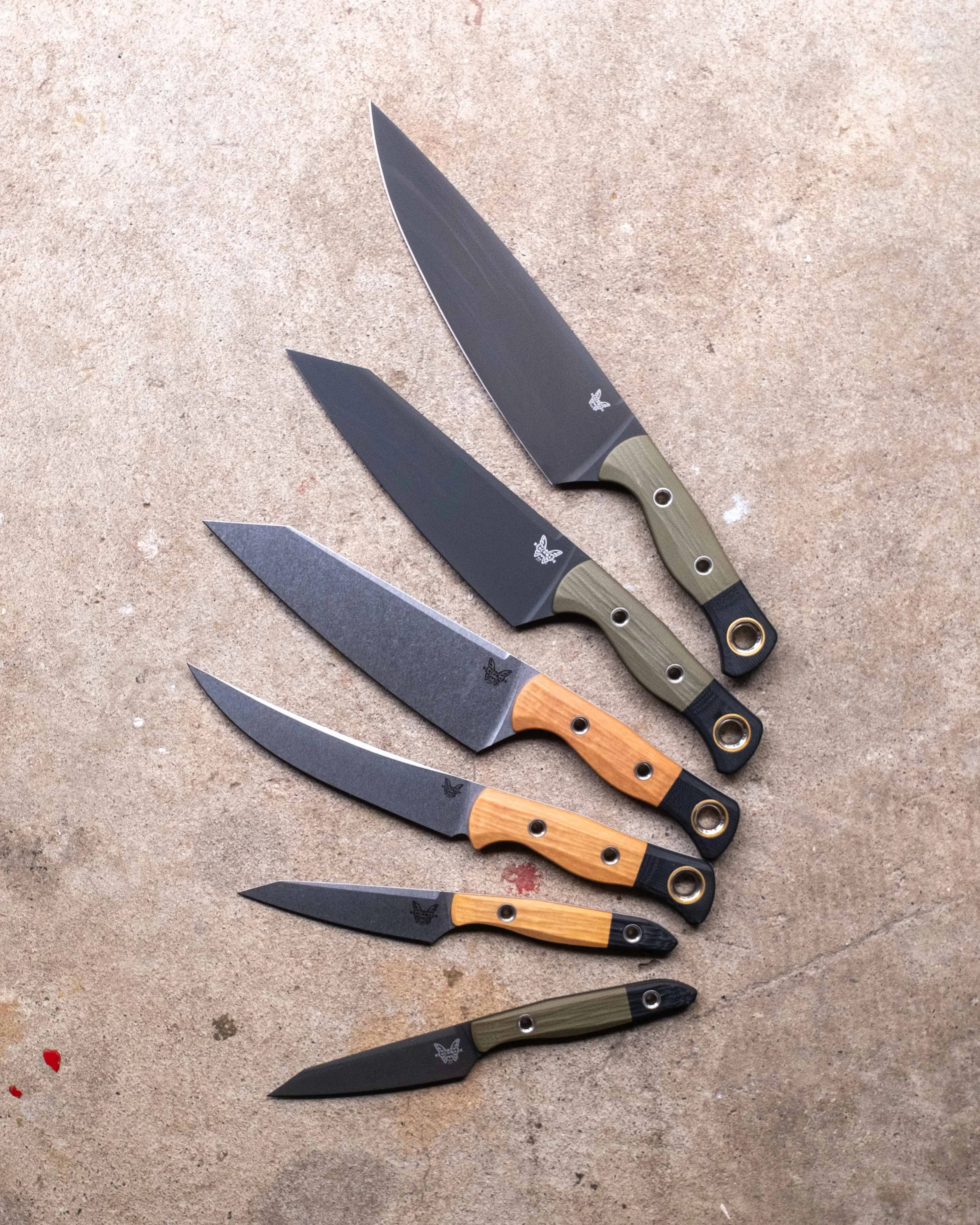 Six kitchen knives with black blades and mixed green and wooden handles arranged in descending size on a concrete surface.