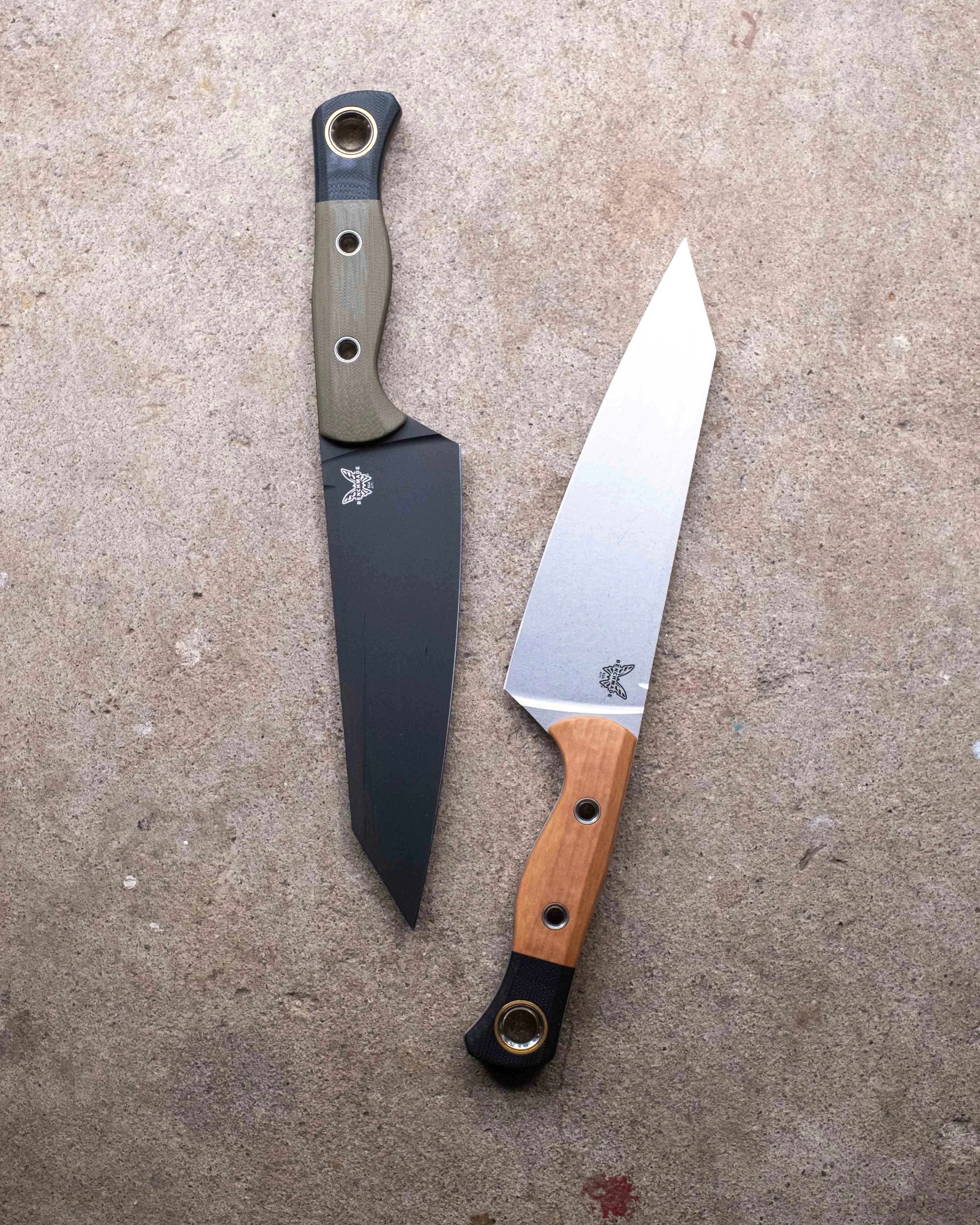 Two fixed-blade knives with angular blades, one with a black blade and green handle, the other with a silver blade and wooden handle, on a concrete surface.