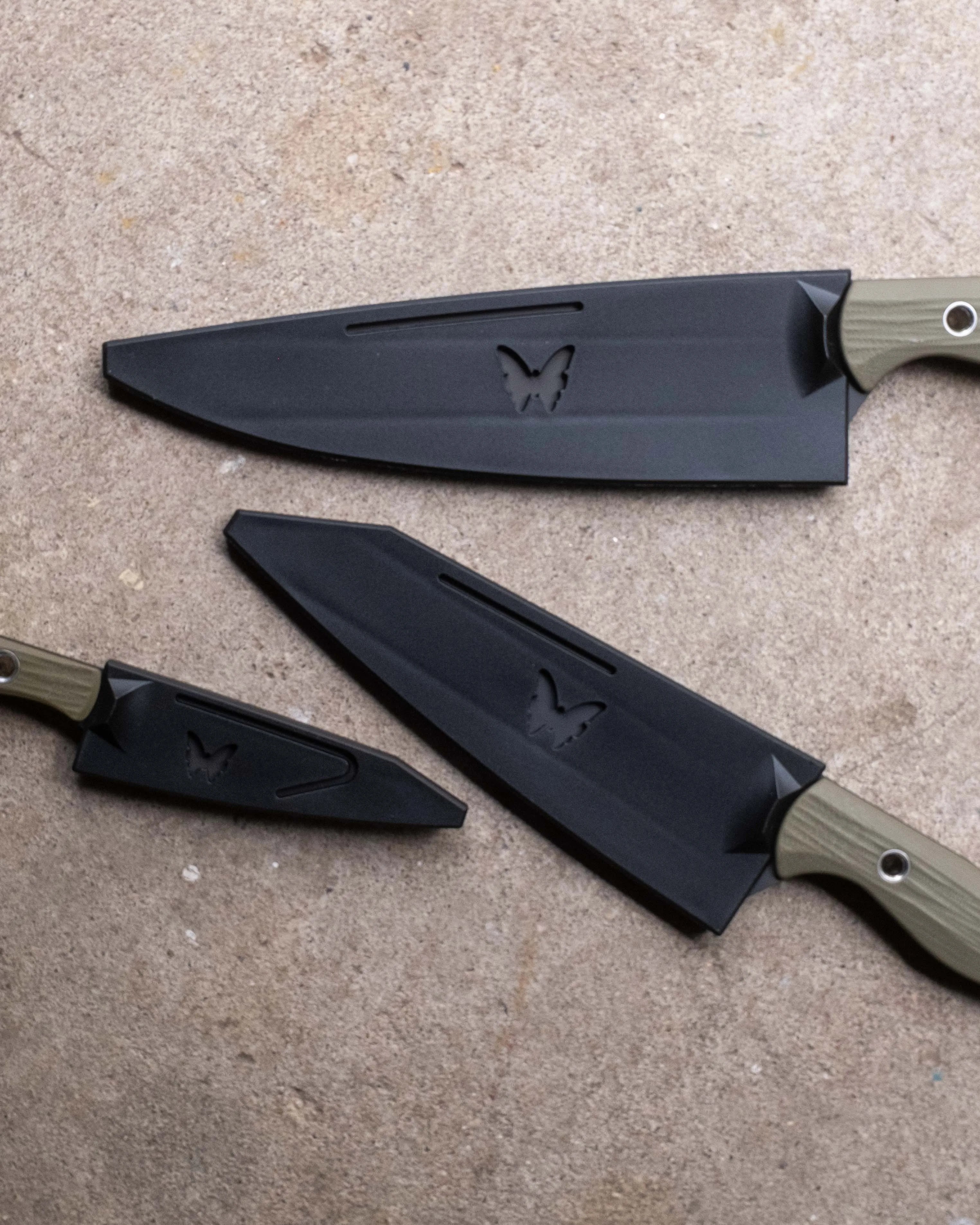 Three black knives with olive green handles and butterfly logos on the blades, laid on a textured surface.